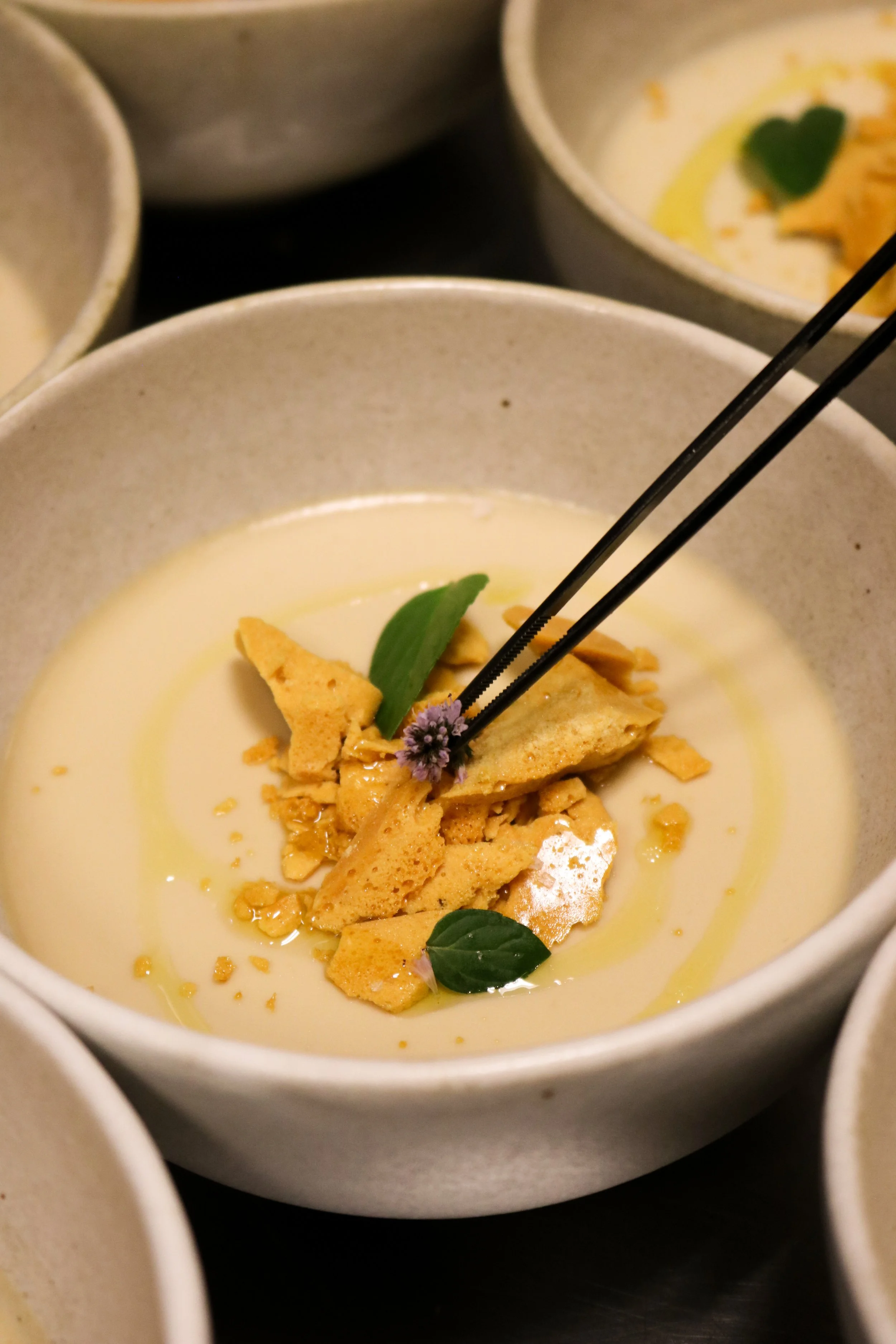 Close-up of a bowl with a creamy yellow soup, garnished with crumbled crackers, a green leaf, and a small purple flower.