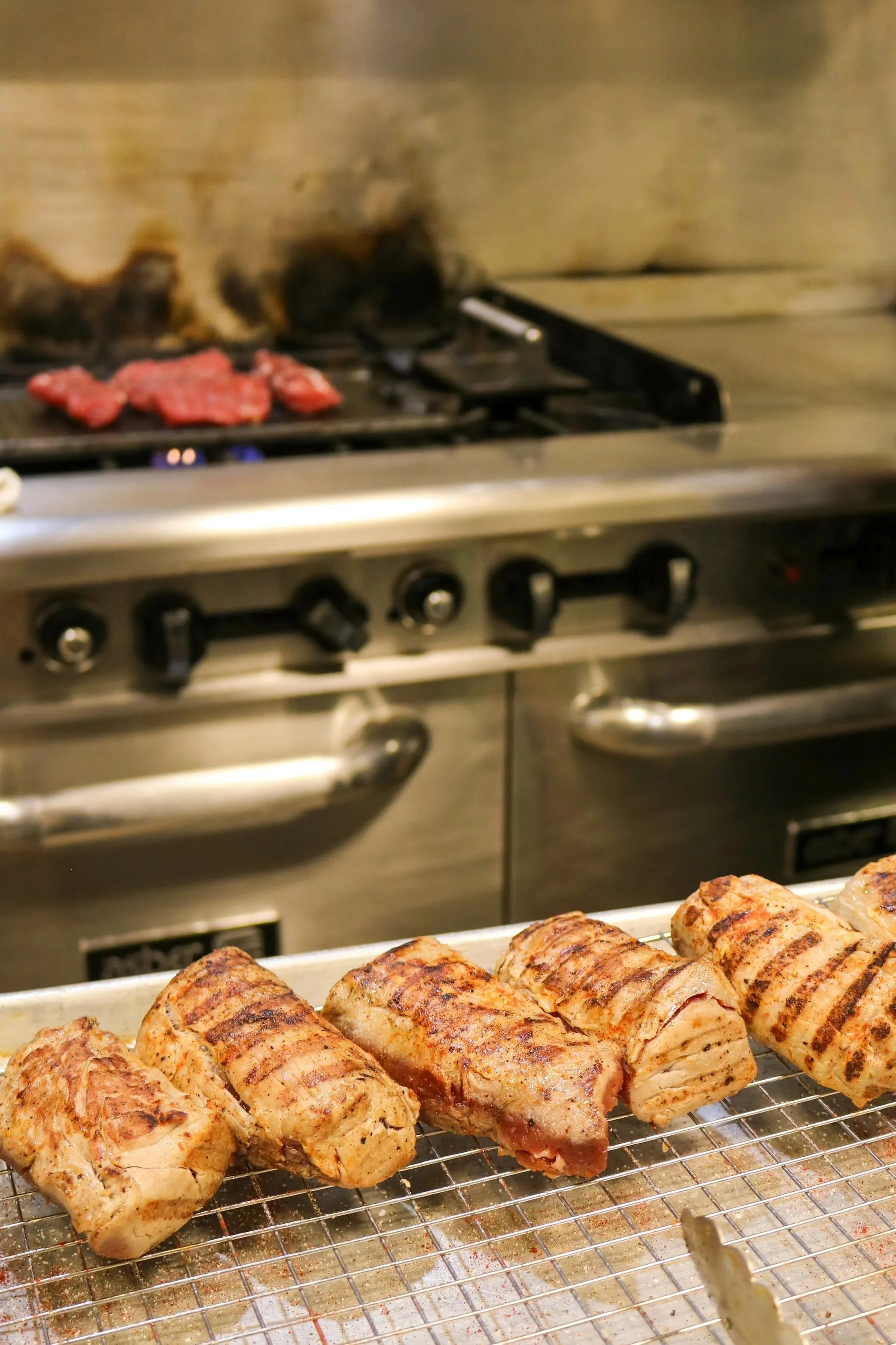 Grilled bacon-wrapped meatloaf rolls on a wire rack in a kitchen oven, with more food in the background.