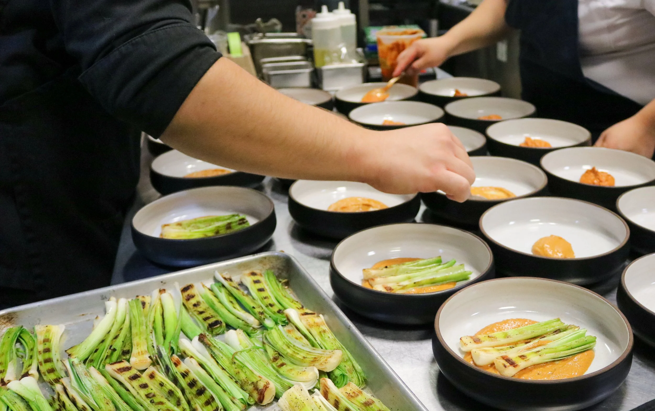 People preparing bowls of sauce with chopped green onions and grilled vegetables or meat in a kitchen.