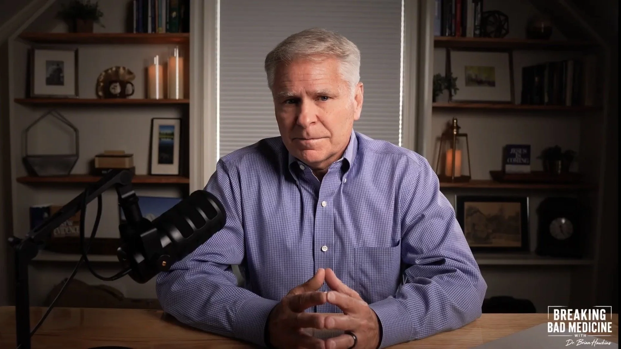 A middle-aged man with gray hair, wearing a blue checkered shirt, sits at a wooden desk with a microphone in front of him. He appears to be speaking or recording. Behind him are wooden shelves with books, framed pictures, candles, and decorative items in a well-lit room. There is a window with blinds behind him, and a small logo in the bottom right corner that reads "Breaking Bad Medicine with Dr. Brian Hawkins."