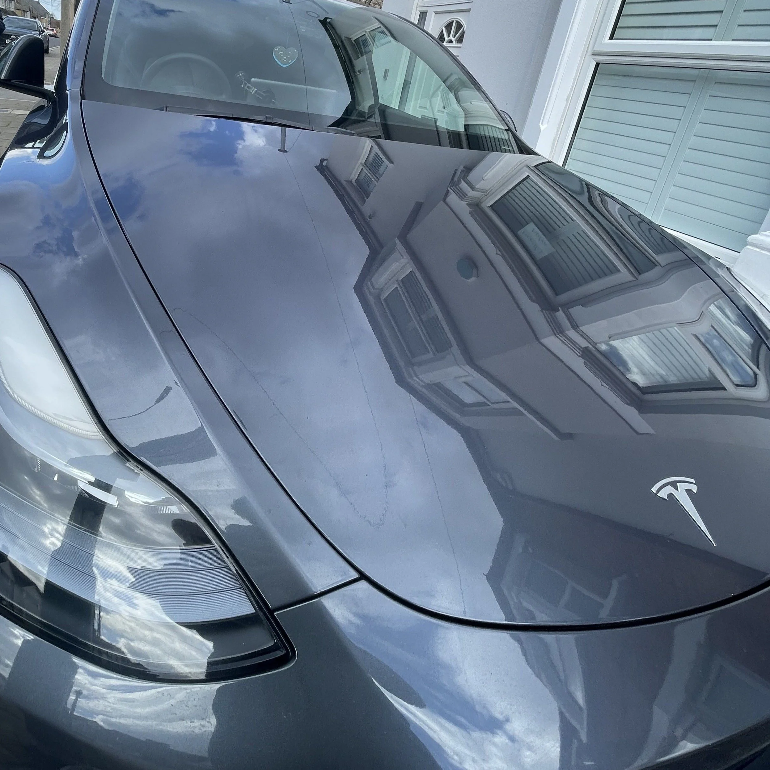 Front view of a dark gray Tesla electric car parked in a driveway with a house and garage in the background, reflecting the sky and surrounding structures on its hood and windshield.
