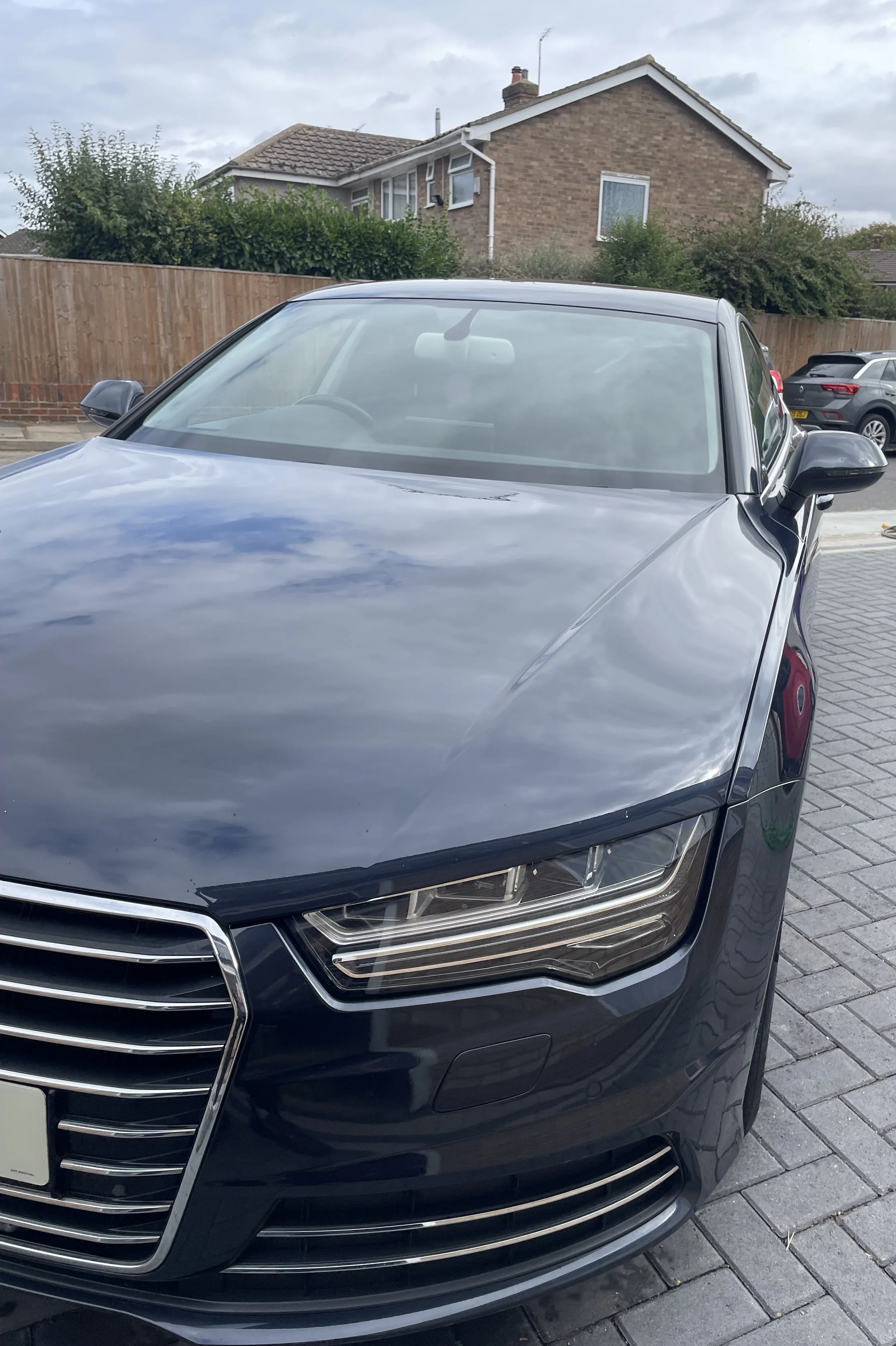 Front view of a gray sedan car parked on a driveway with residential houses and a brick fence in the background.