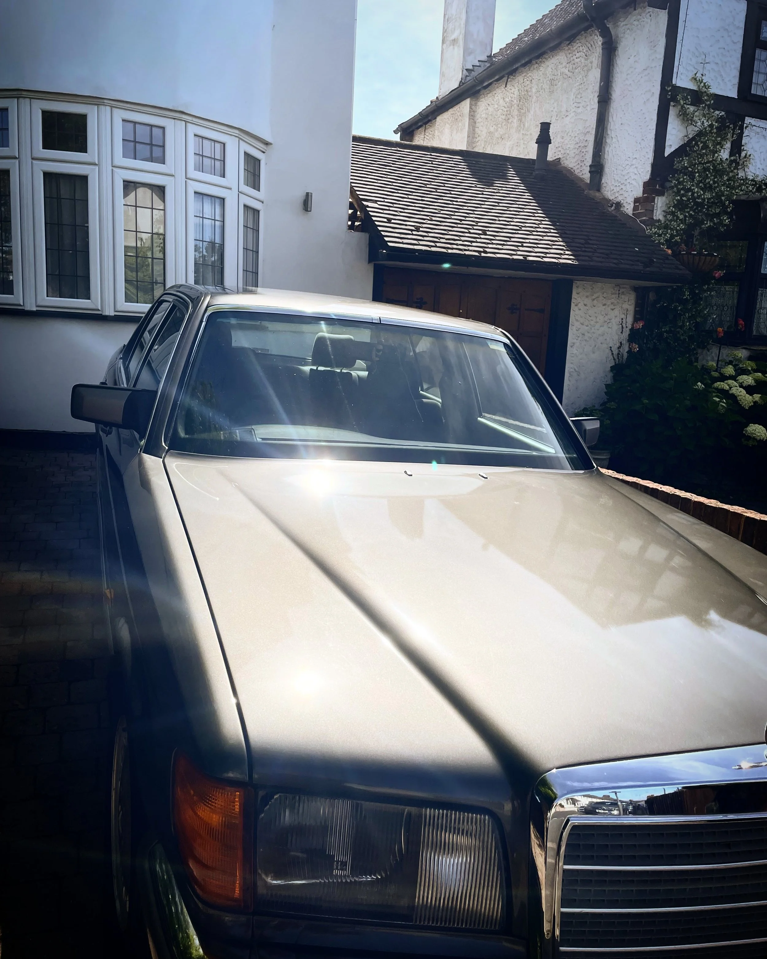 Close-up of the front of a silver car parked outside a house. The house has a bay window with multiple panes and a white exterior. The roof of the house has shingles, and there are some plants and flowers near the house.