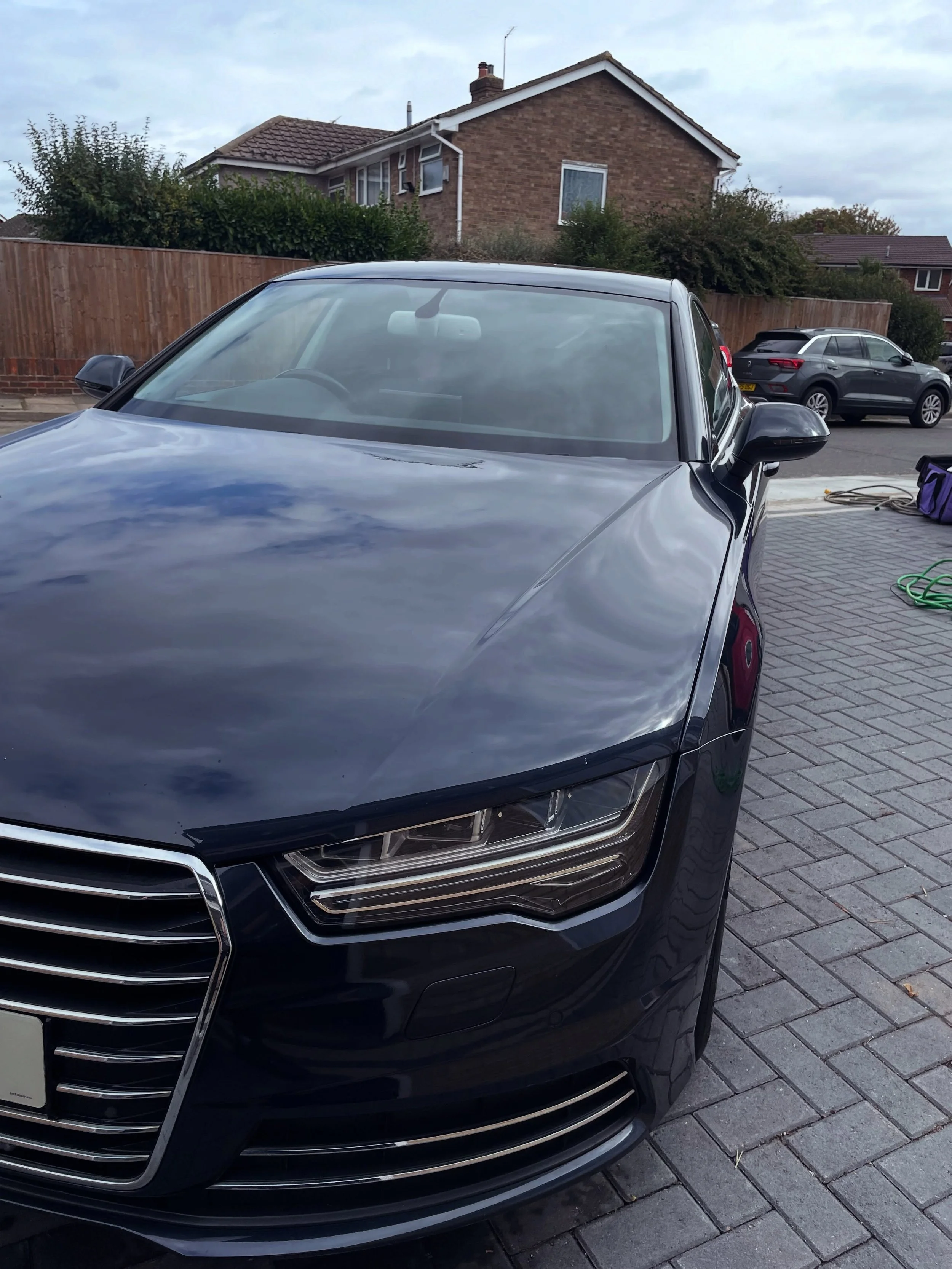 A black sedan parked on a brick driveway in front of a residential house with a wooden fence. There is another car parked behind it on the street and garden hoses are on the ground.