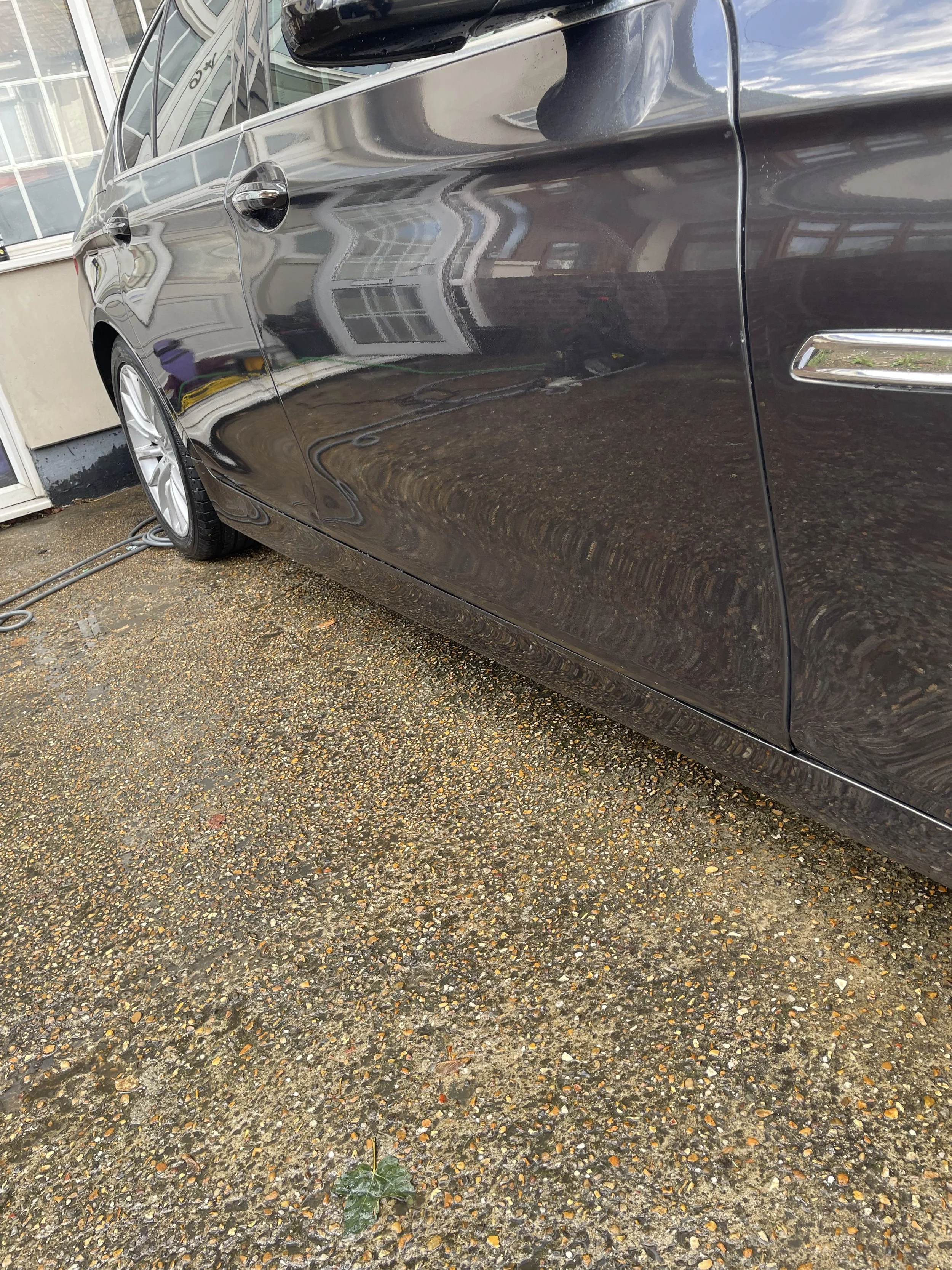 Black car parked on gravel driveway next to building with large windows, reflections of the building visible on car surface, small green plant on ground in foreground.