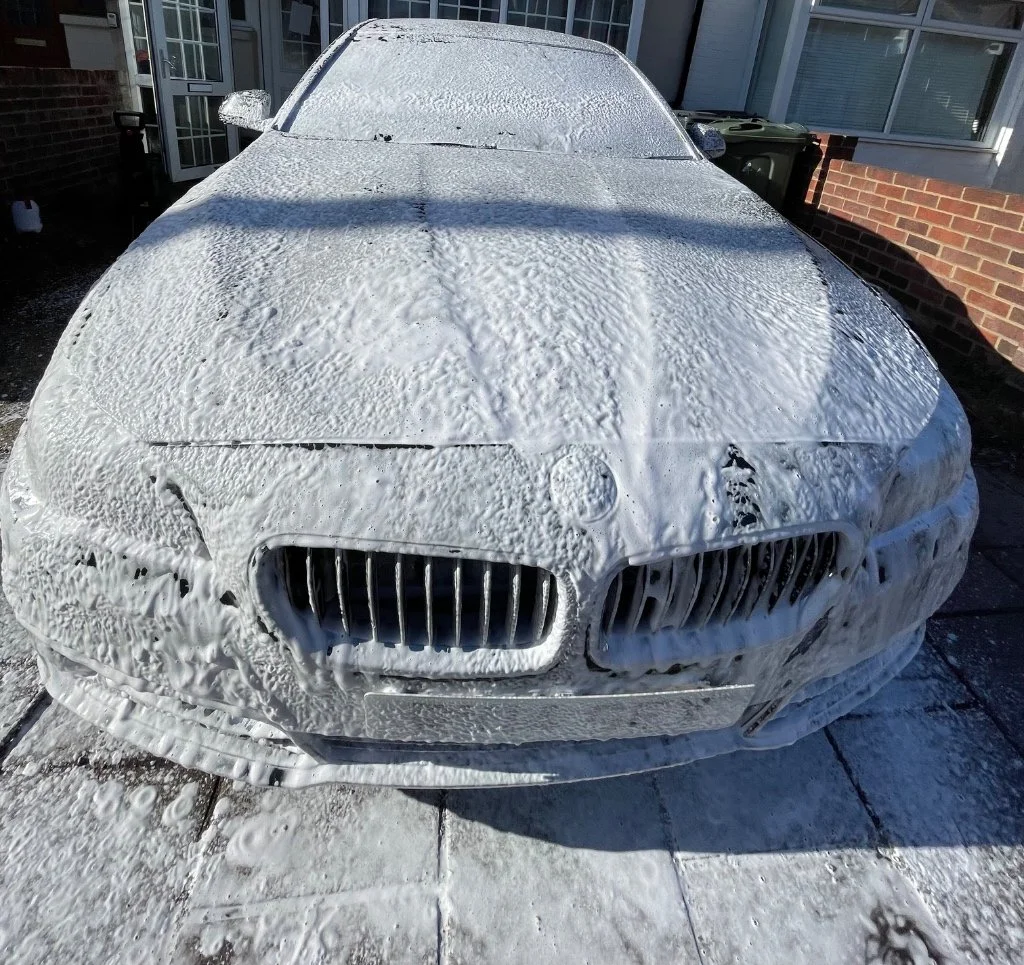 A car covered in soap suds and foam, parked outside a house with brick and siding walls, during daytime.