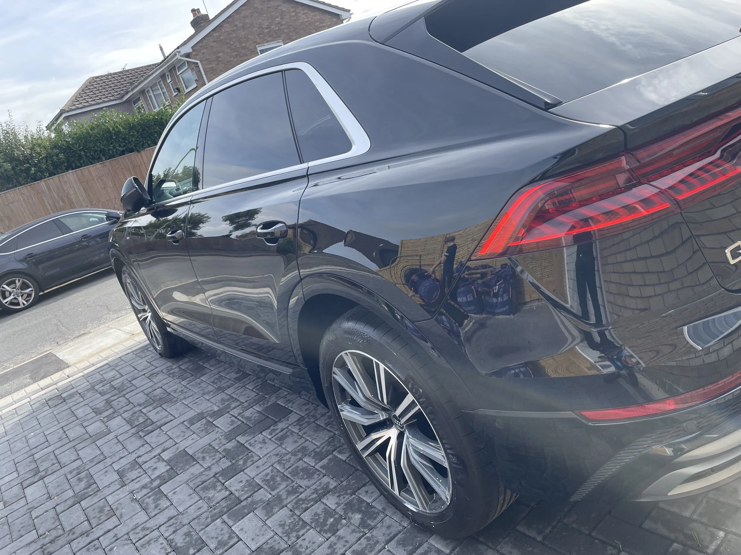 A shiny black luxury SUV parked on a paved driveway with a wooden fence and residential house in the background.