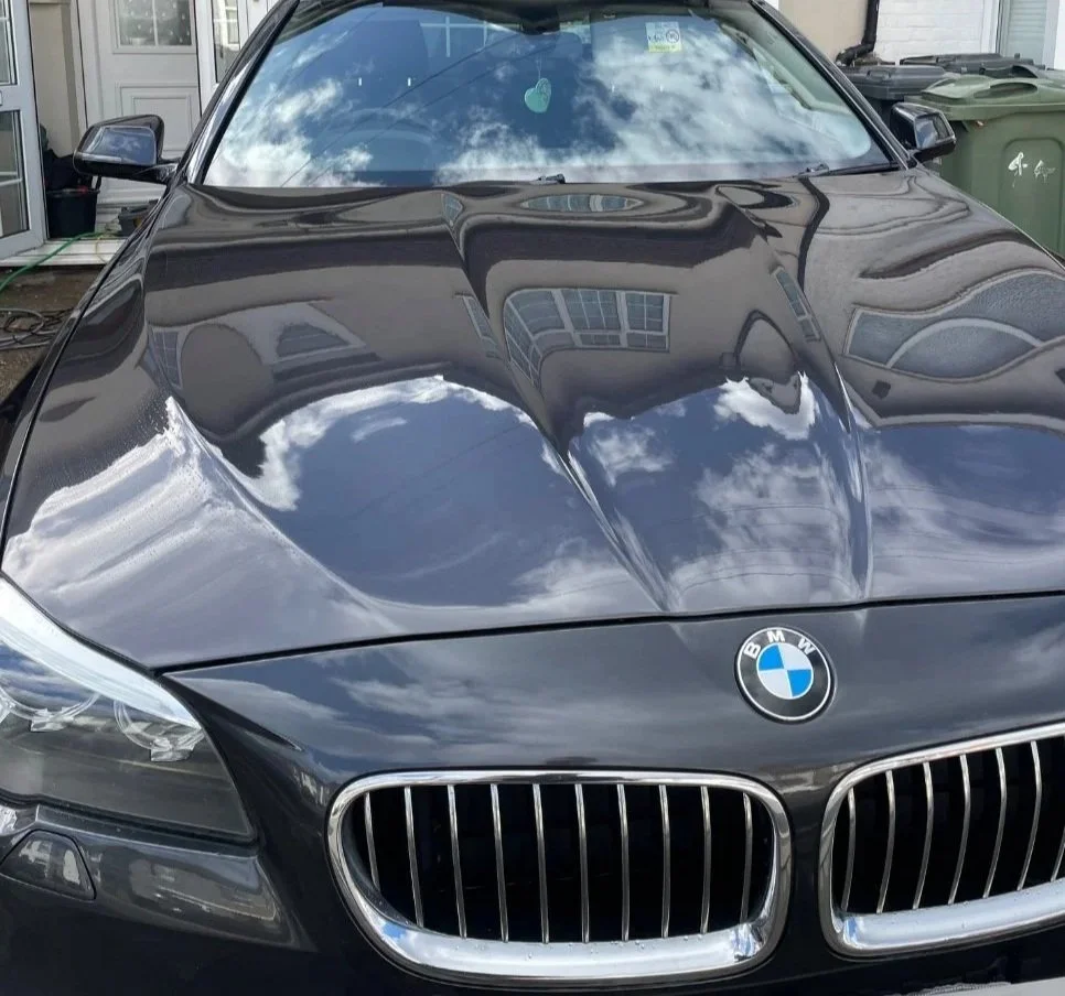 A black BMW car with a reflective hood showing the sky and clouds, parked in a driveway next to some green trash bins.