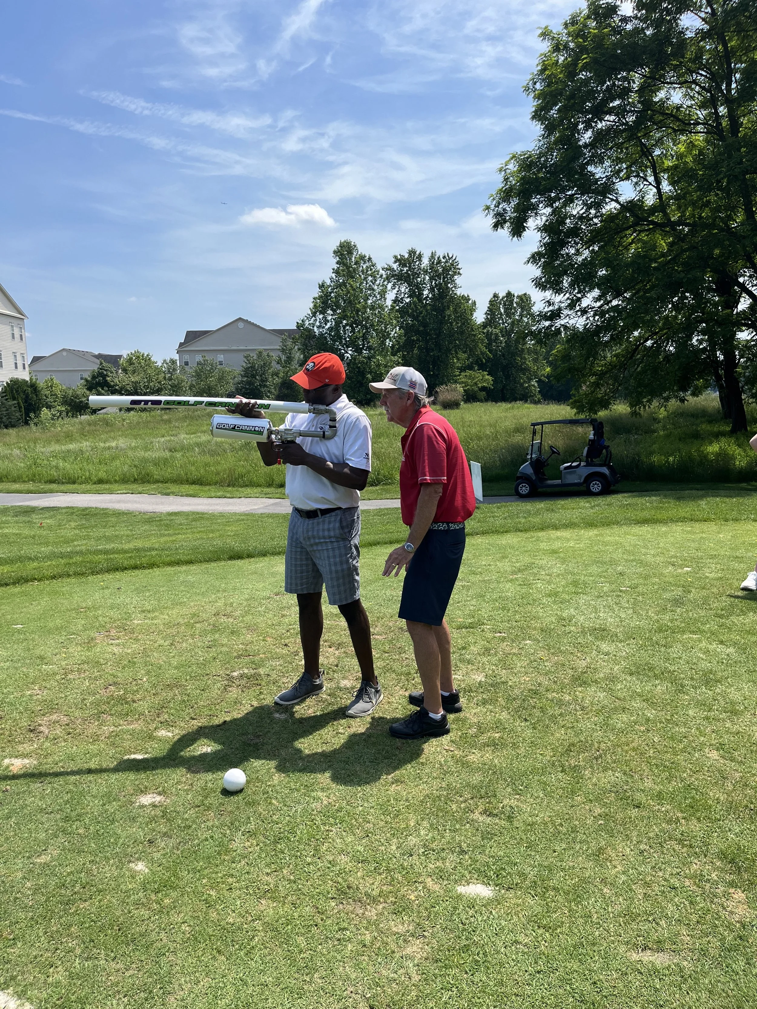 Two men on a grassy golf course with a Golf Cannon launcher, golf cart, and ball under a sunny sky