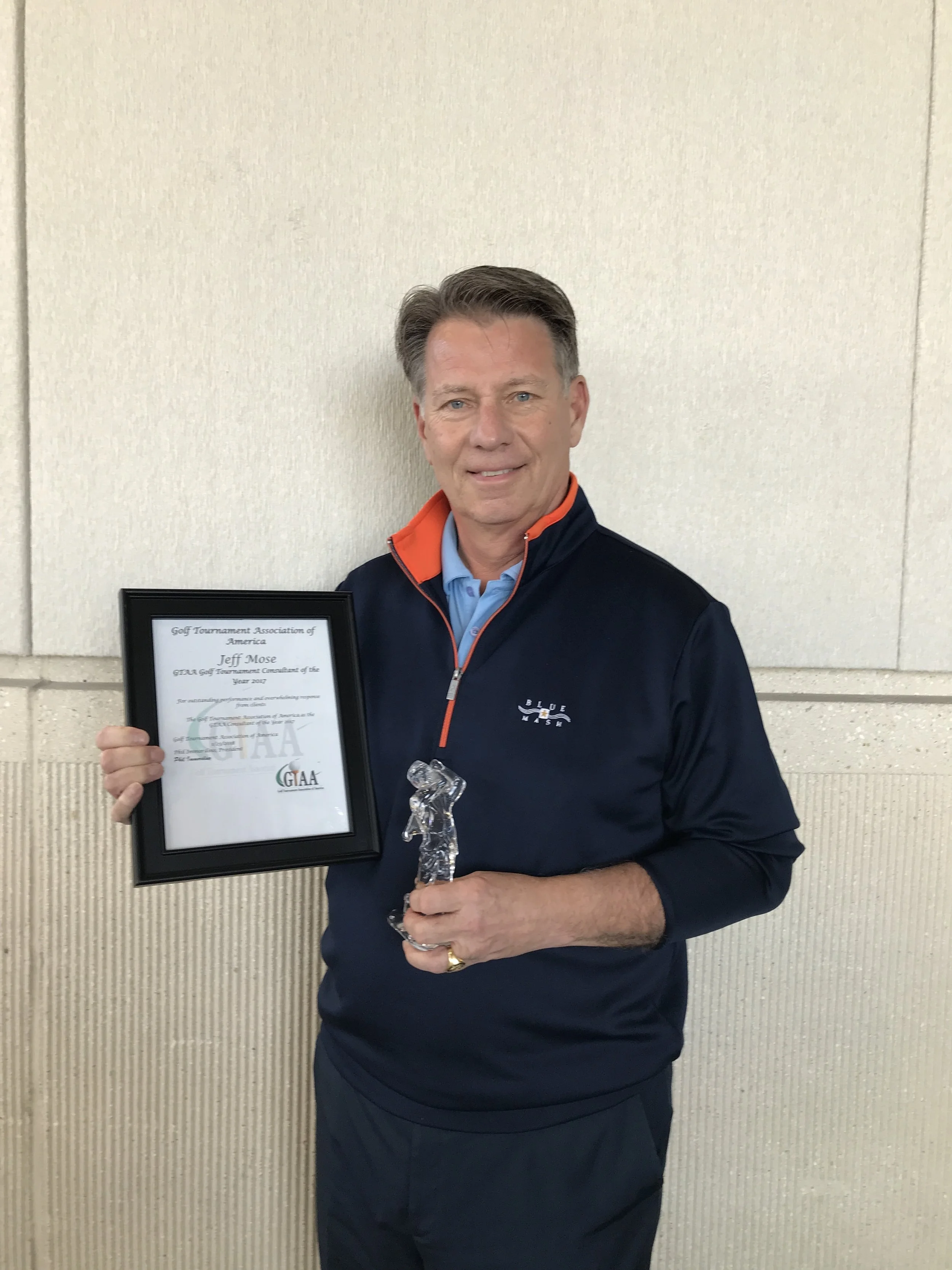 Man holding a framed Golf Tournament Association of America award and crystal golf trophy