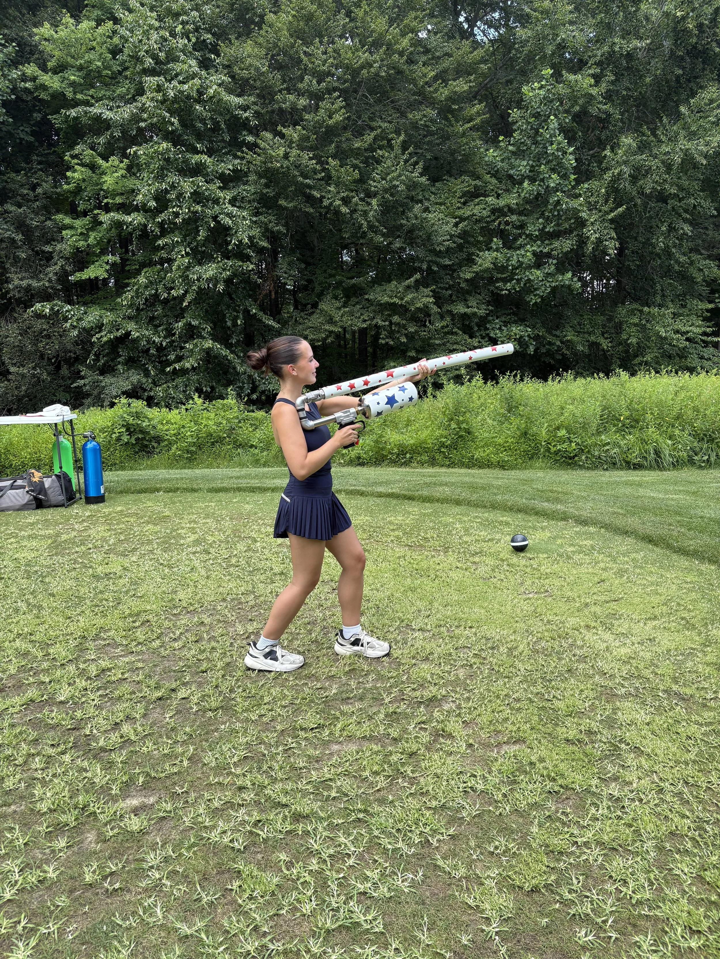 Woman in a pleated athletic skirt holding a patriotic star-patterned T-shirt cannon on a grassy field, with trees in the background.