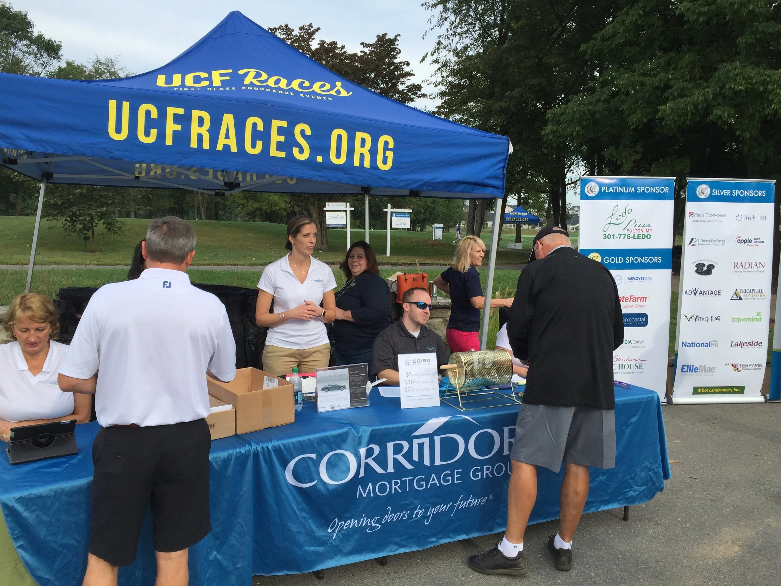 UCF Races charity event booth with volunteers, sponsor banners, raffle drum, and registration table outdoors