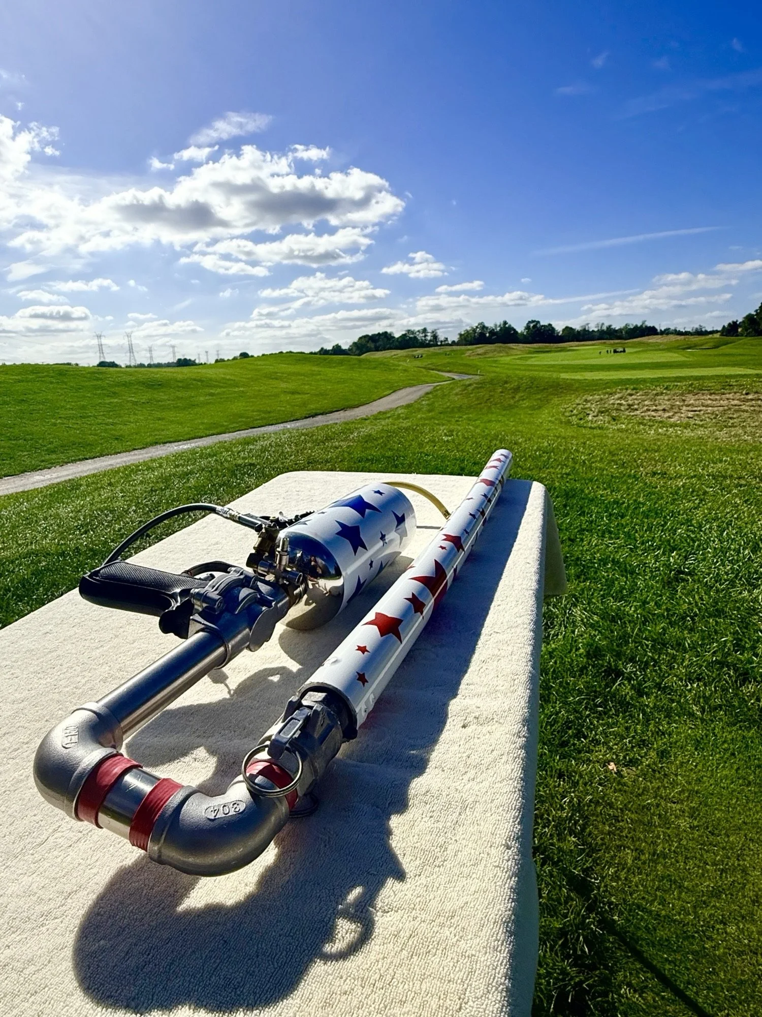 Patriotic red, white, and blue paintball guns resting on a towel beside a grassy field under a blue sky
