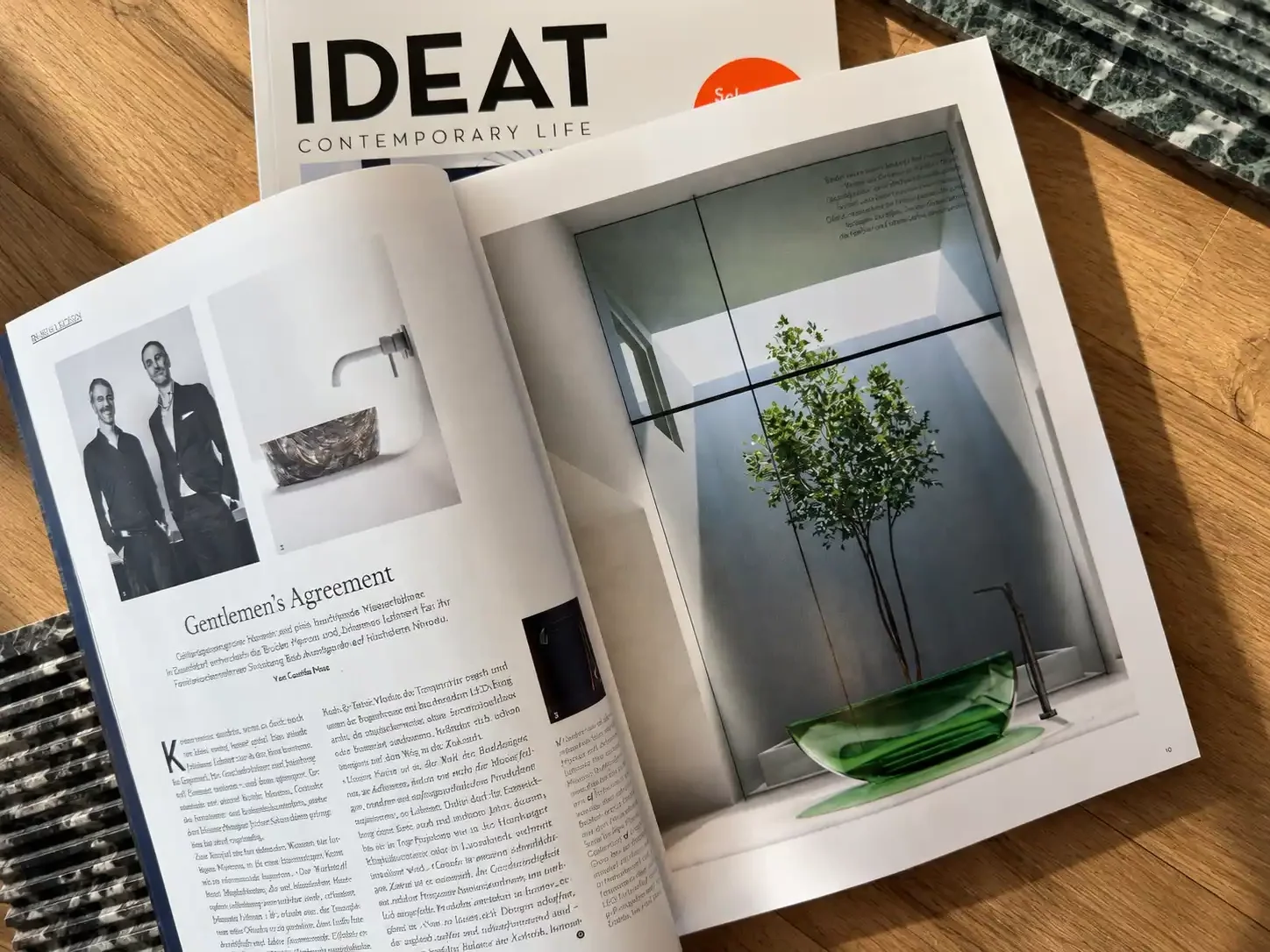 Open magazine with black-and-white photos of two men in suits, a modern sink, and an article titled 'Gentlemen's Agreement'. Next page shows a minimalist interior with a small tree in a green glass planter by a window.