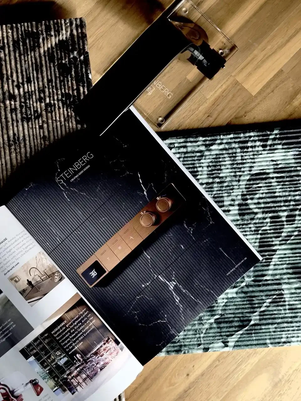 Open magazine on a wooden surface showing a brown digital controls panel and black marble pattern beside a black and white patterned cloth. A transparent box with the word "STEINBERG" and a metallic box are also nearby.