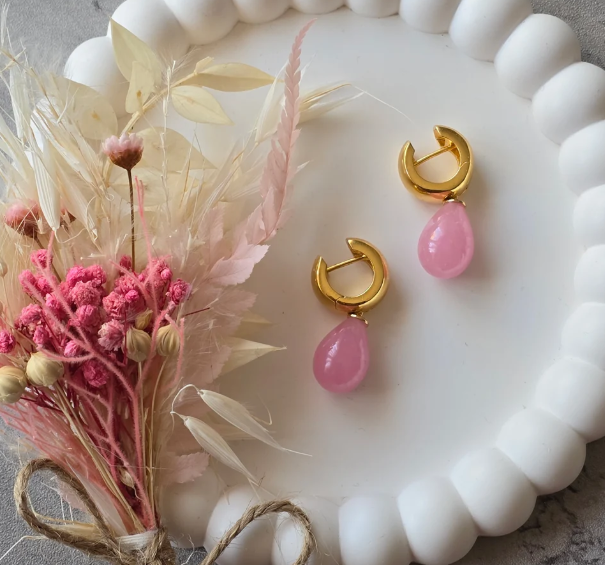 Ohrringe mit goldenen Kreisen und pinken Tropfen auf einem weißen Porzellanservice, daneben getrocknete Blumen in Pastellfarben.