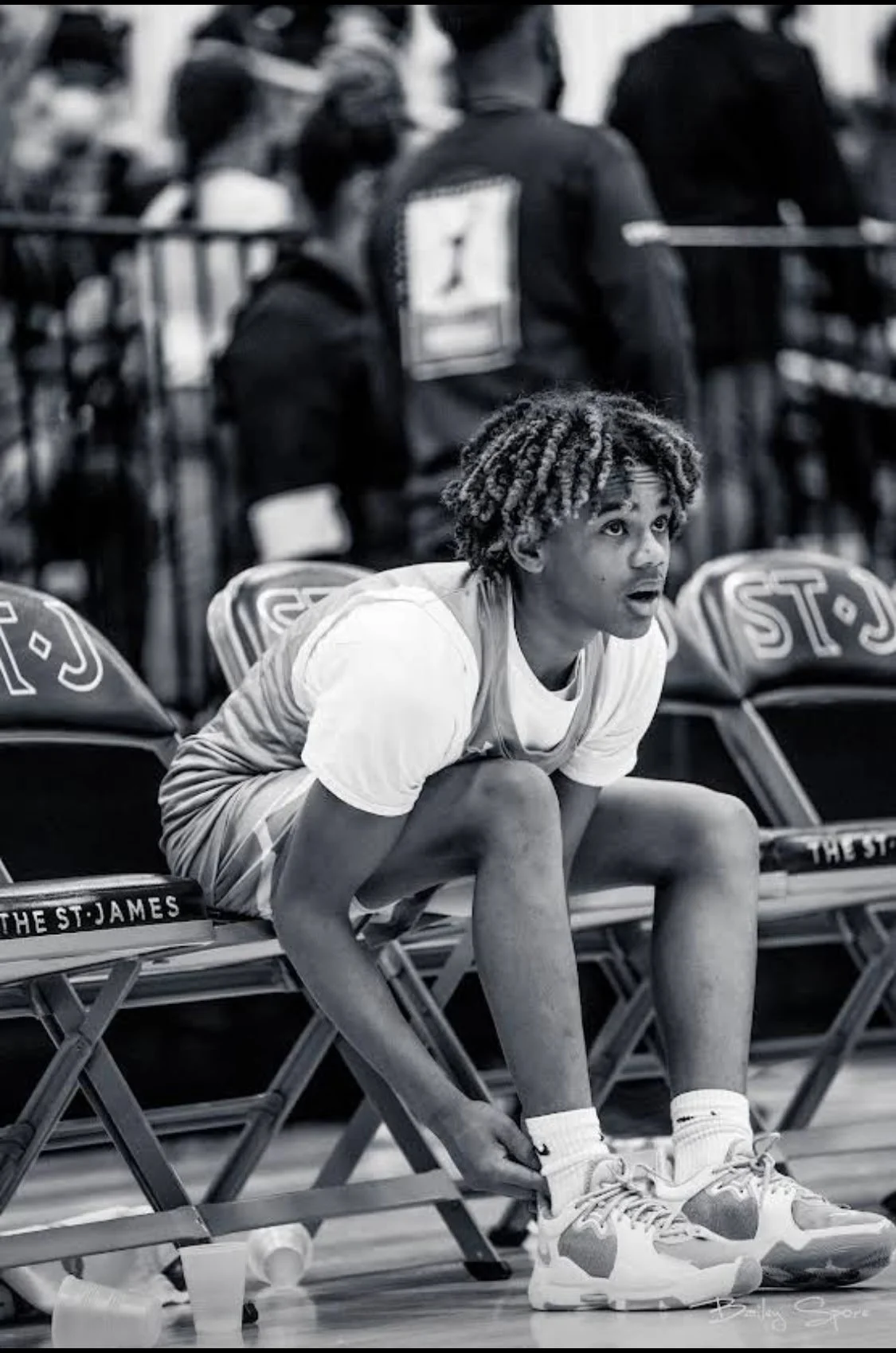 A young basketball player with dreadlocks sits on a bench, leaning forward and tying his shoelaces during a game or practice. He is wearing a white t-shirt and basketball shoes, with the bench and empty chairs behind him.