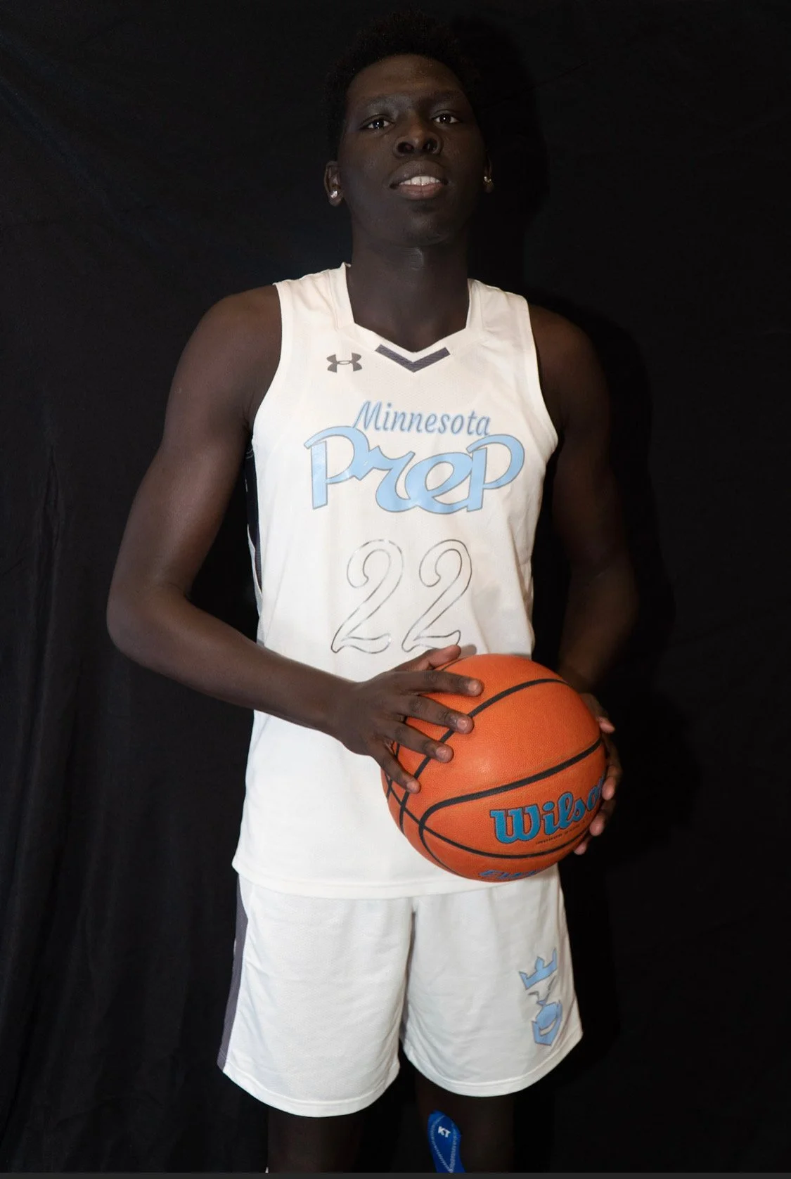 A female basketball player in a white Minnesota Prep basketball uniform holding an orange Wilson basketball, standing against a black background.