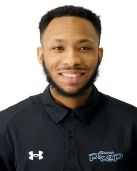A young man with a beard and short curly hair, smiling, wearing a black collared shirt with a logo that says 'Track and Field Prep'.
