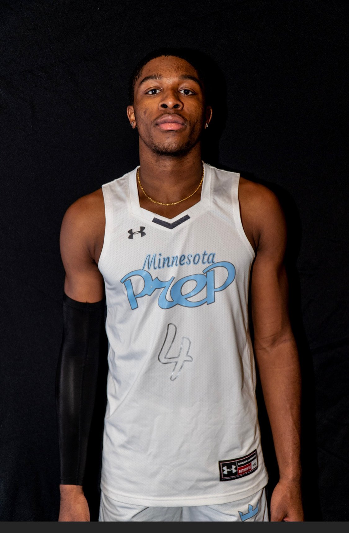 Young male basketball player in a white Minnesota Prep jersey with number 4, black sleeve on his right arm, standing against a black background.