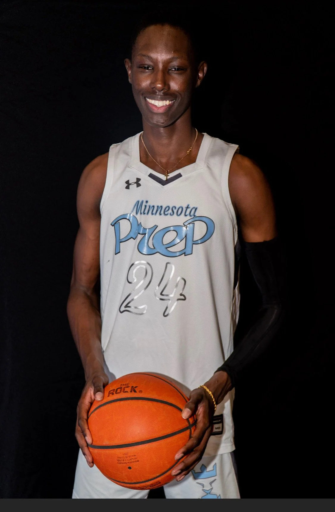 A young man wearing a white Minnesota Prep basketball jersey with the number 24, holding an orange basketball, smiling at the camera against a black background.