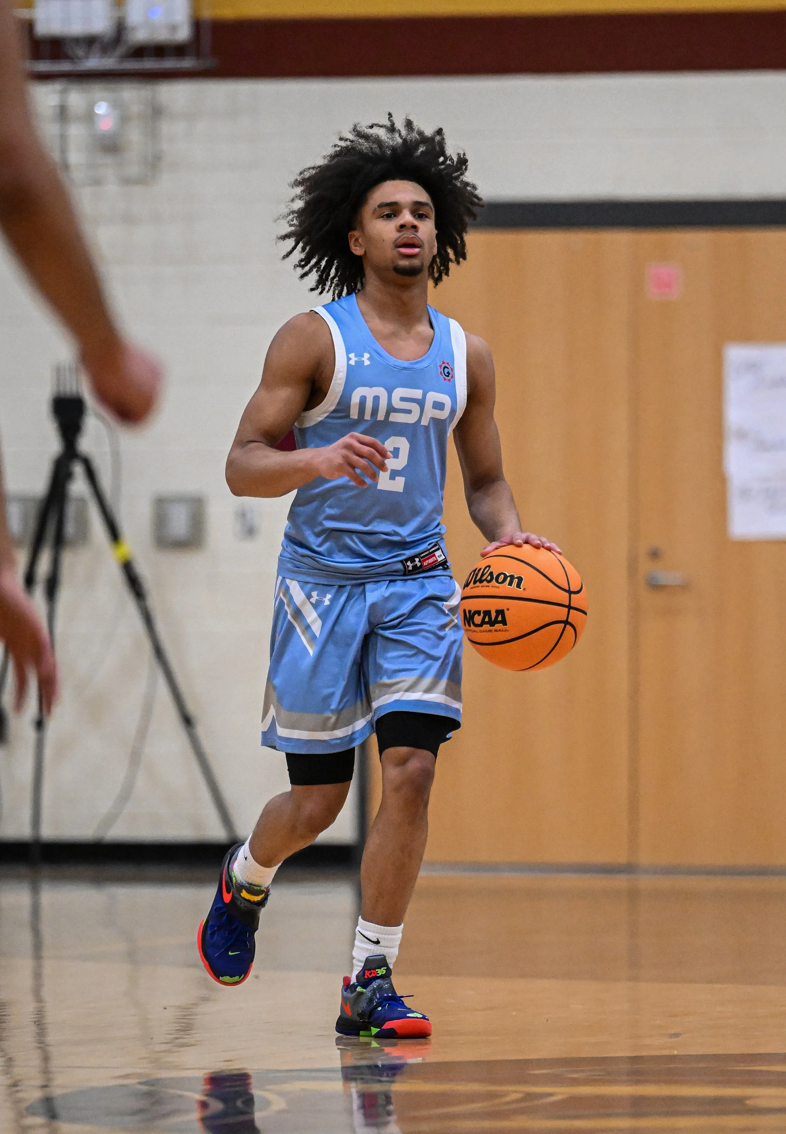 A young male basketball player with curly hair, wearing a blue jersey with 'MSP' and the number 2, dribbling a Wilson basketball on an indoor court.