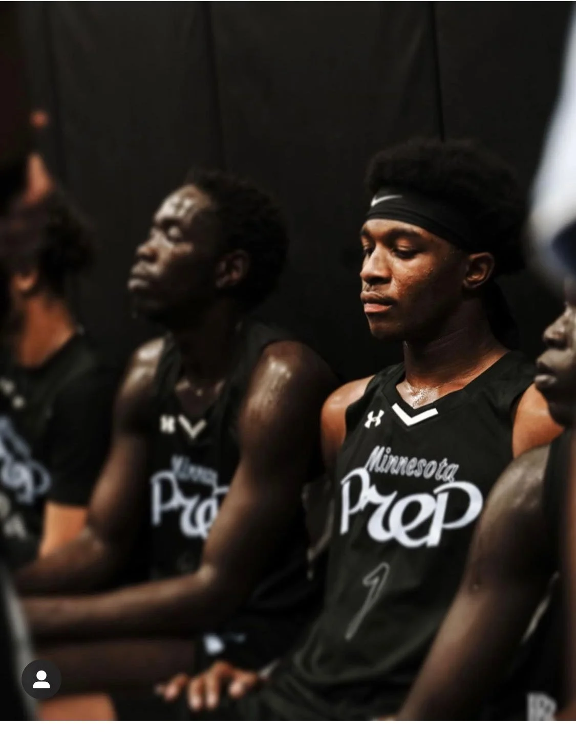 A group of young men in black Minnesota Prep basketball uniforms are sitting with eyes closed, appearing to meditate or focus. They are seated in a row against a dark background, with serious and calm expressions.