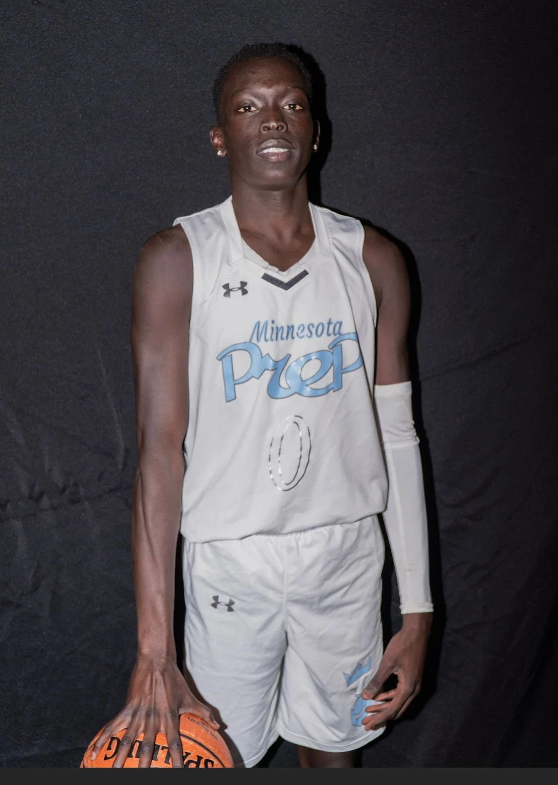 A female basketball player in white Minnesota Prep jersey number 0, holding an orange basketball, standing against a dark background.
