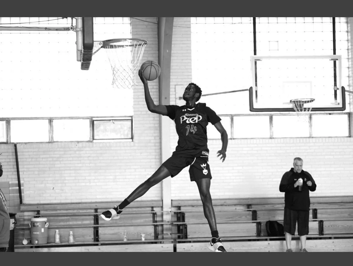 A basketball player in a black jersey with the number 14 is jumping to make a dunk in an indoor basketball court. A coach or spectator is standing on the sideline with a water bottle.