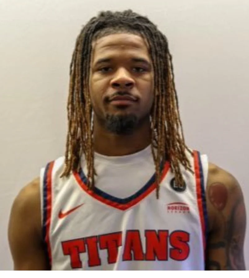 A young man with dreadlocks wearing a white basketball jersey that says 'Titans' and a small Nike logo, standing against a plain gray wall.