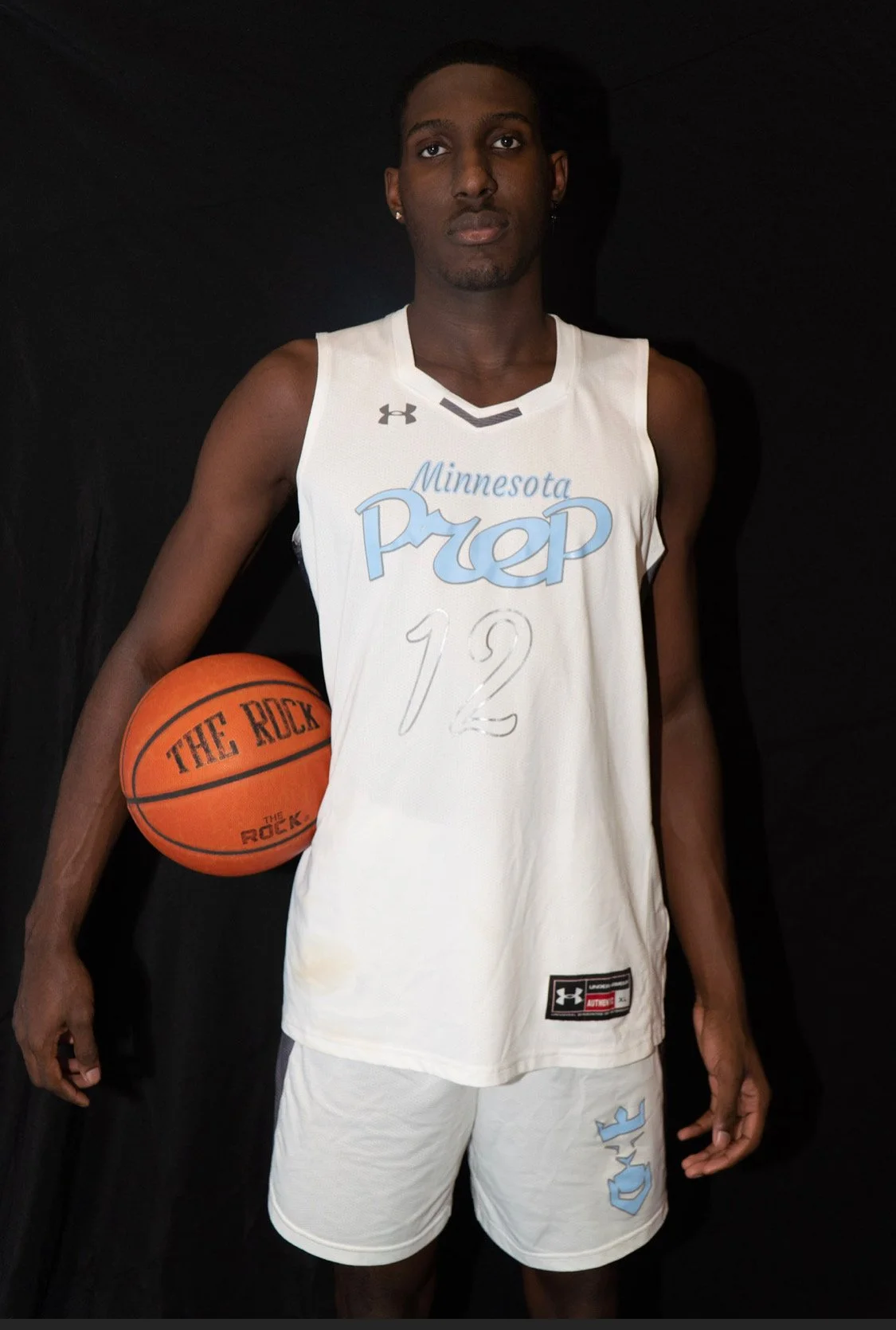 A young African American male basketball player in a white jersey with ''Minnesota Prep'' and the number 12, holding an orange basketball marked ''The Rock'', standing against a black background.