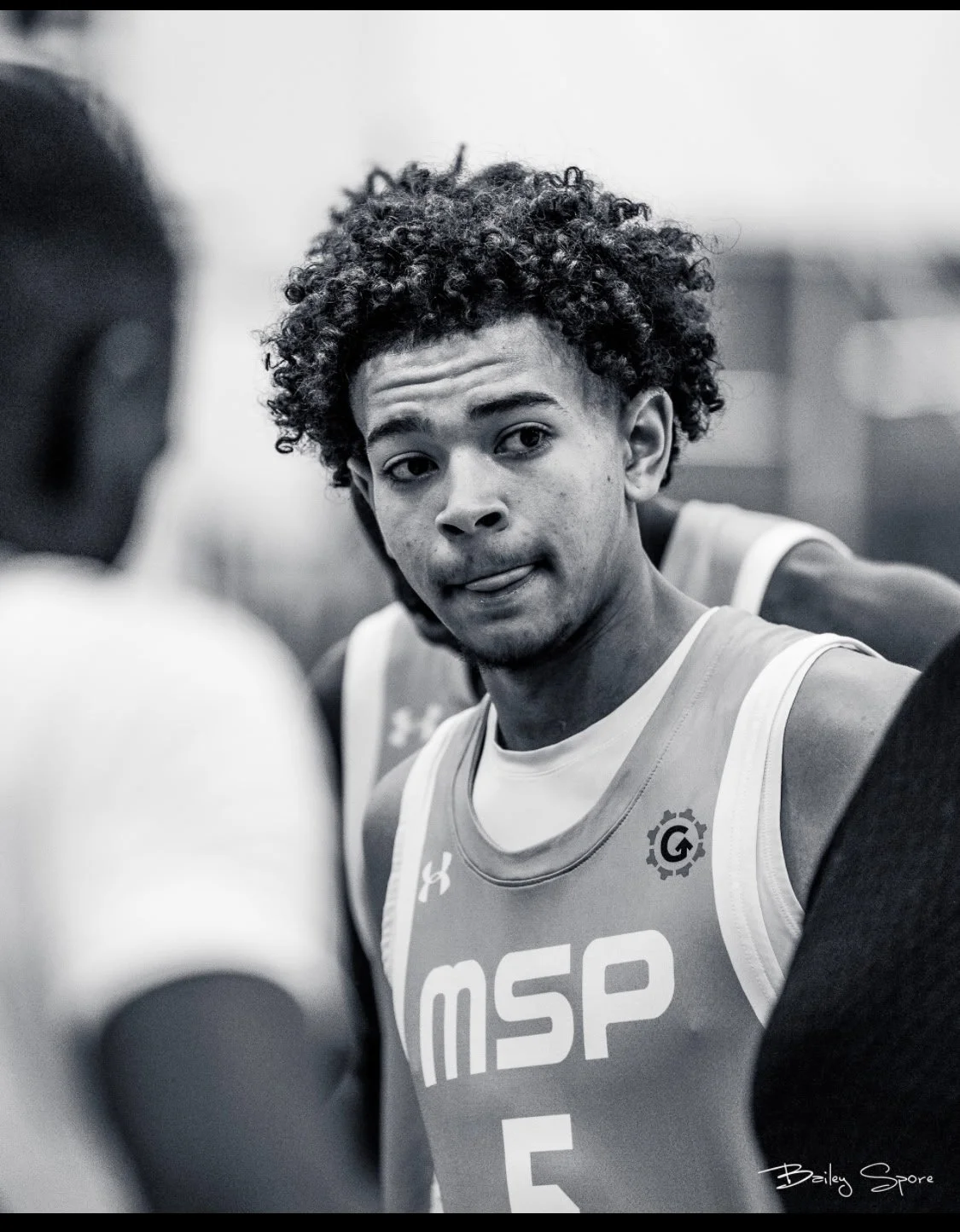 Black and white photo of a young male basketball player with curly hair, wearing a sports jersey, looking at the camera with a slightly open mouth and visible tongue, in a gym or arena setting.
