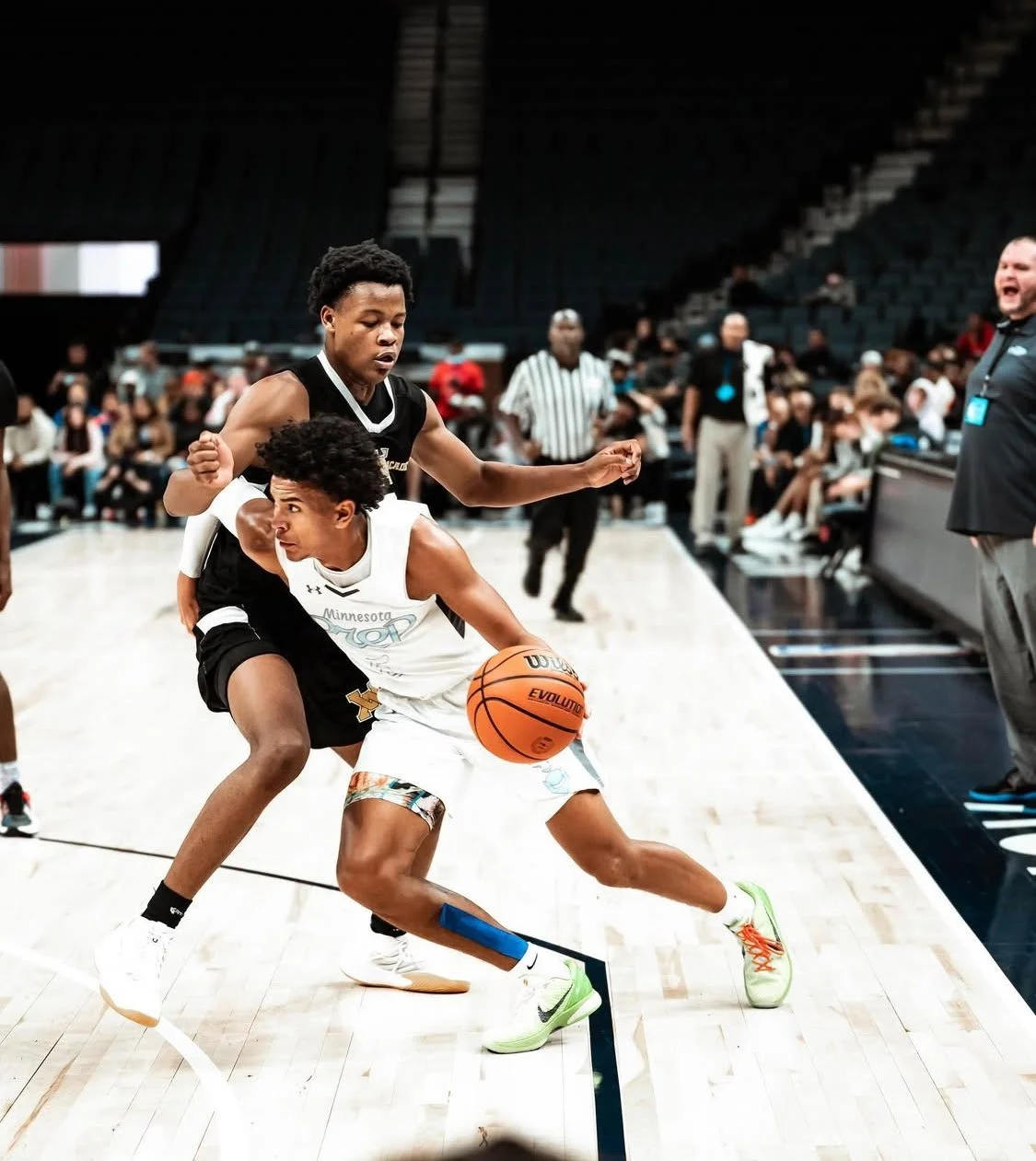 Two young basketball players compete for the ball during a game, with one player in a white jersey and the other in a black jersey. Spectators and officials are watching in the background.