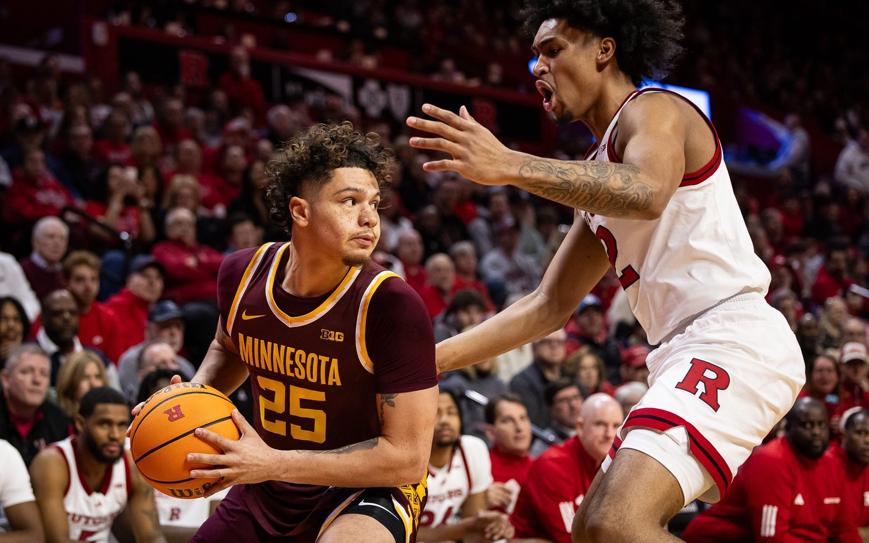 A basketball player from the Minnesota Golden Gophers, wearing a maroon and gold uniform with number 25, is holding a basketball while being guarded closely by a player from Rutgers University, who is wearing a white uniform with red accents. They are playing in a packed basketball arena.