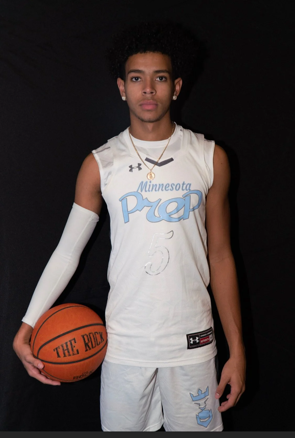 A young man in a white basketball uniform with 'Minnesota Prep' and the number 5, holding a basketball labeled 'The Rock,' standing against a black background.