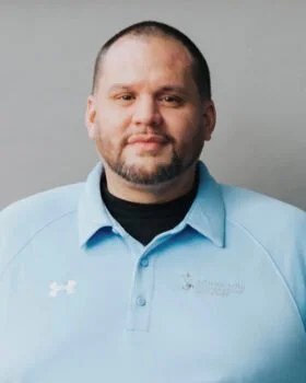 Portrait of a man with short hair and a beard, wearing a light blue collared shirt with logos, against a plain gray background.