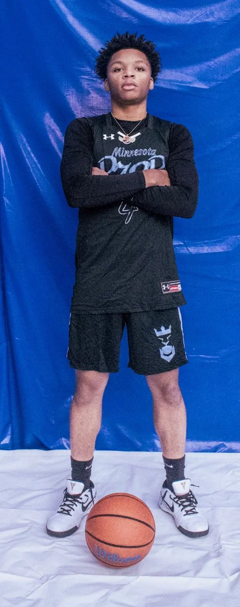 Young adult standing with arms crossed behind a basketball on a white surface, wearing a black Minnesota Valor sports jersey and shorts, white high-top sneakers, black socks, and a silver chain necklace, against a blue backdrop.