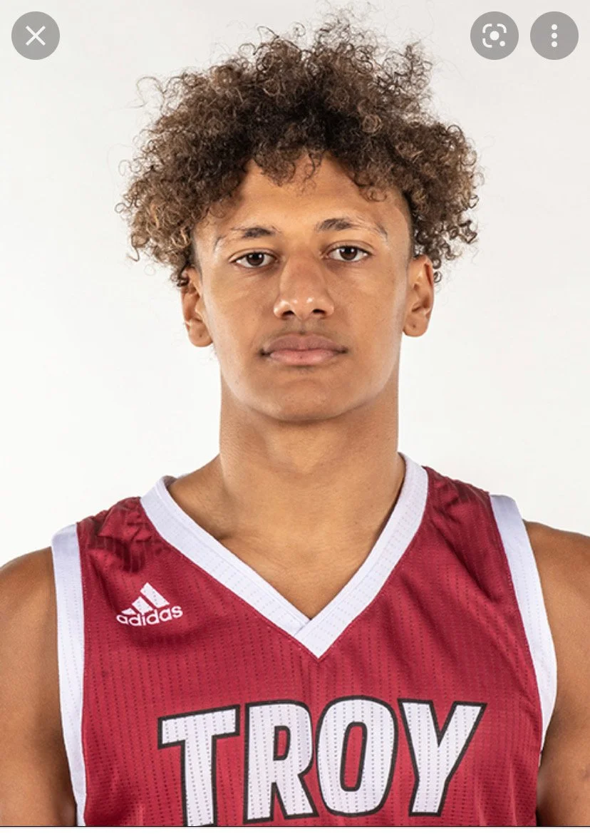A young man with curly brown hair, wearing a red basketball jersey with white trim and the word 'TROY' on the front, standing against a plain white background.