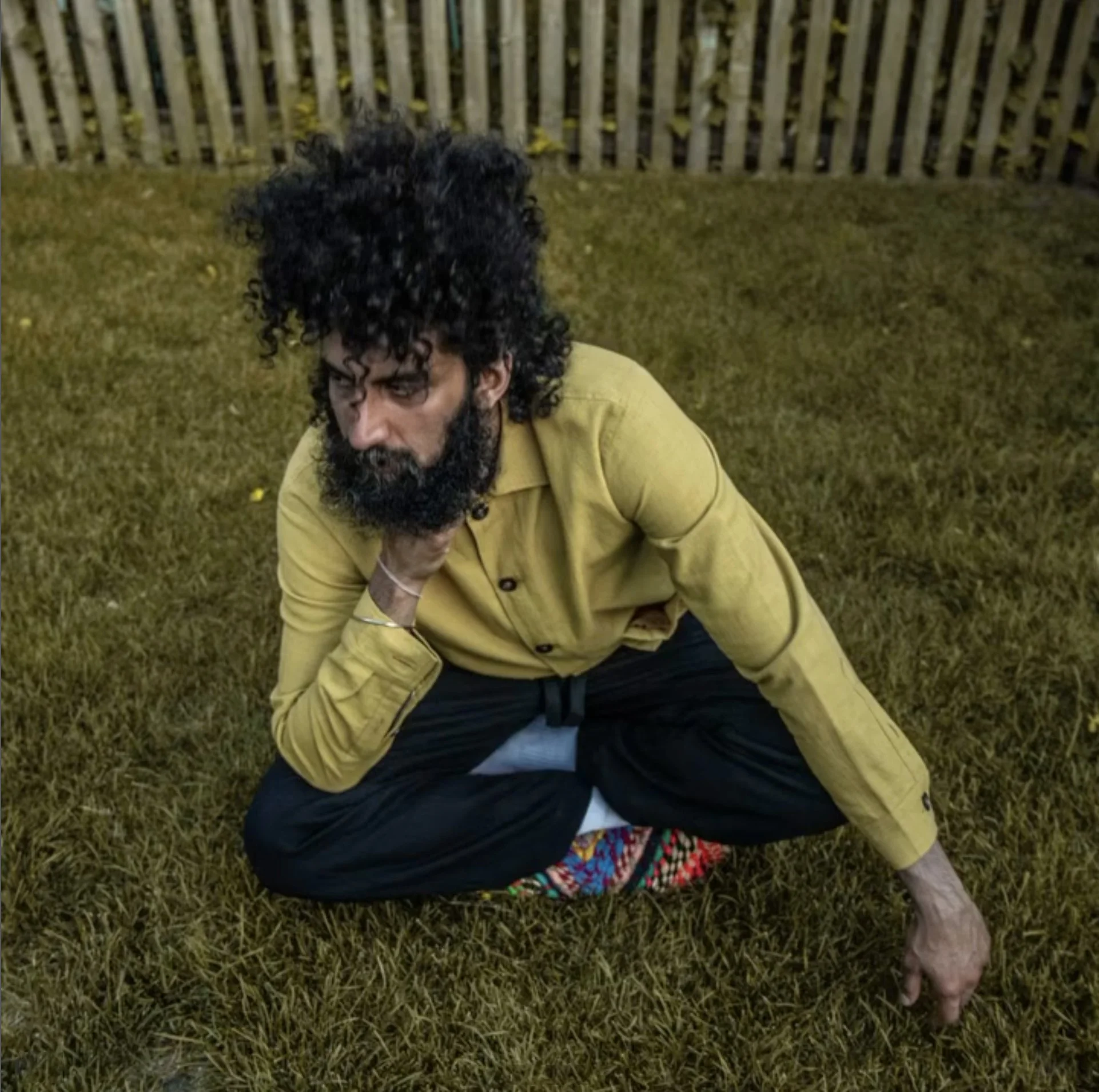 A person with long curly hair and a beard, wearing a yellow jacket, sitting on grass with a fence in the background.