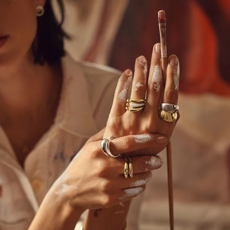 A person's hands covered in white paint, holding a thin paintbrush vertically with fingers adorned with multiple rings.