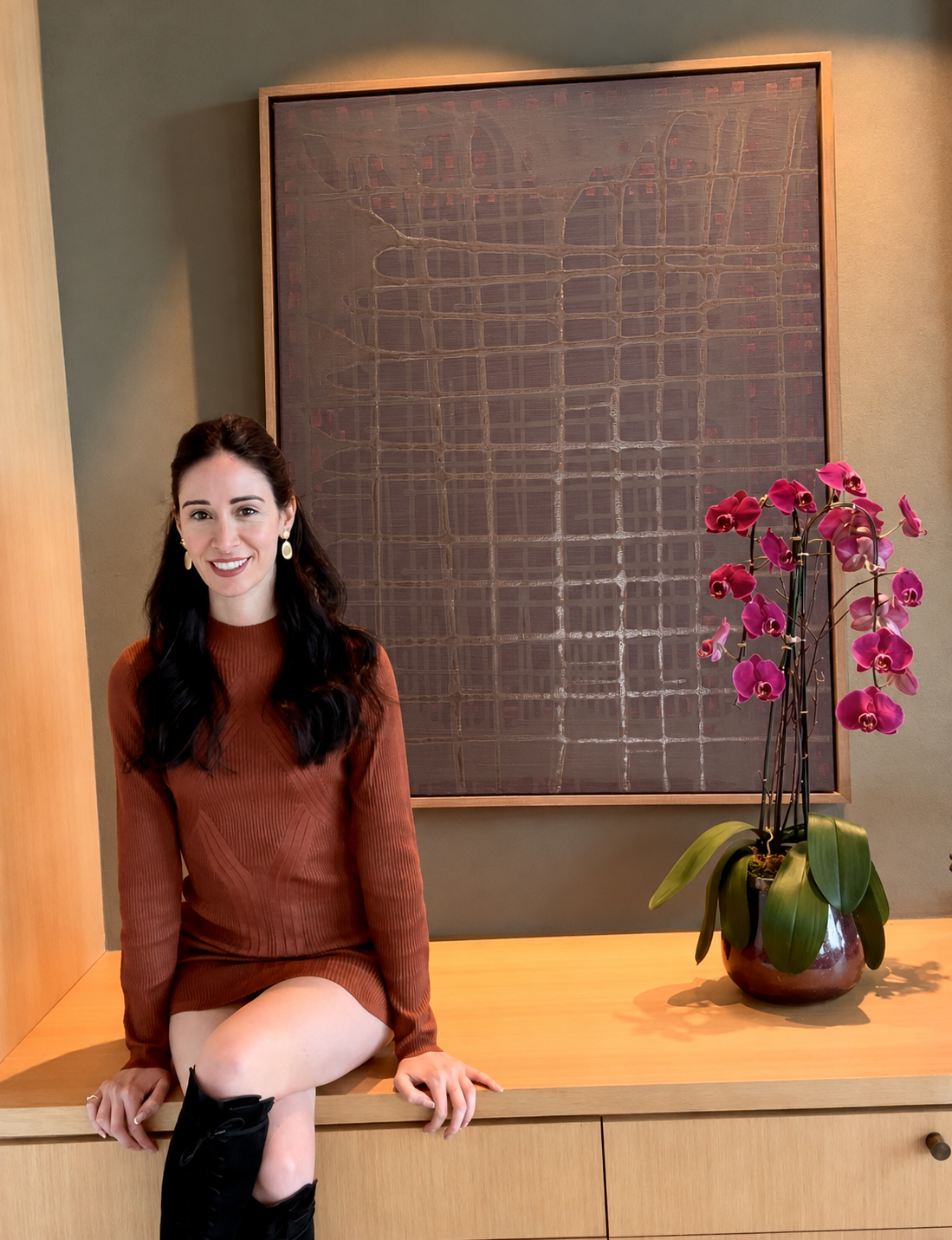 A woman with long dark hair sitting on a wooden cabinet, wearing a rust-colored long-sleeve dress and black boots, smiling at the camera. Behind her is a large abstract painting with a brown background and a pink orchid plant in a ceramic pot.