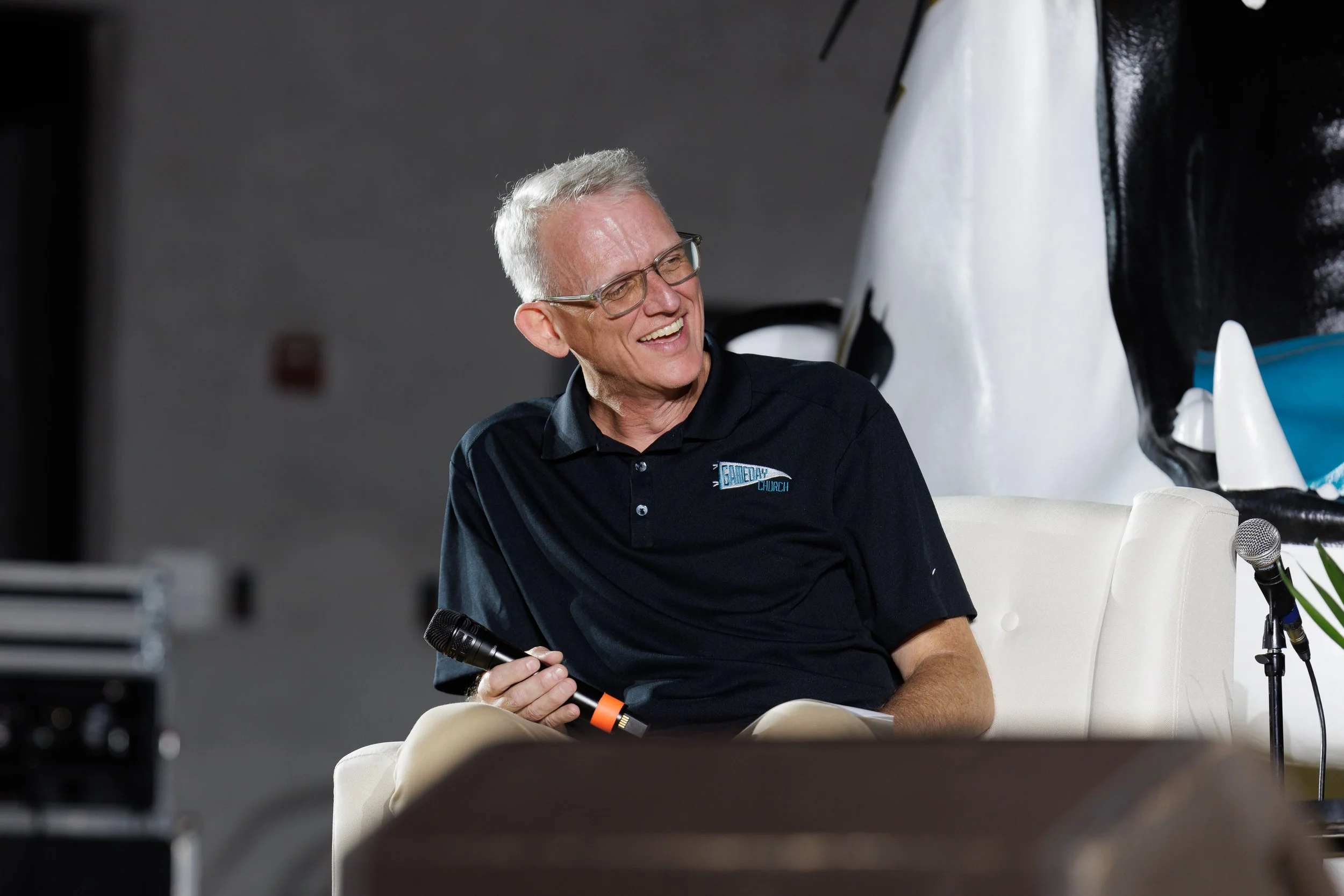 Pastor David Tarkington, smiling and holding a microphone, sitting on a white chair at the Jaguars Faith, Family, & Football event.