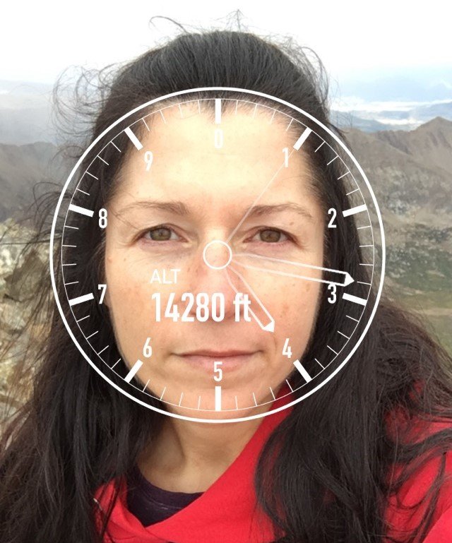 A woman standing outdoors in a mountainous area with a digital overlay of a clock face showing an altitude of 14,280 feet.
