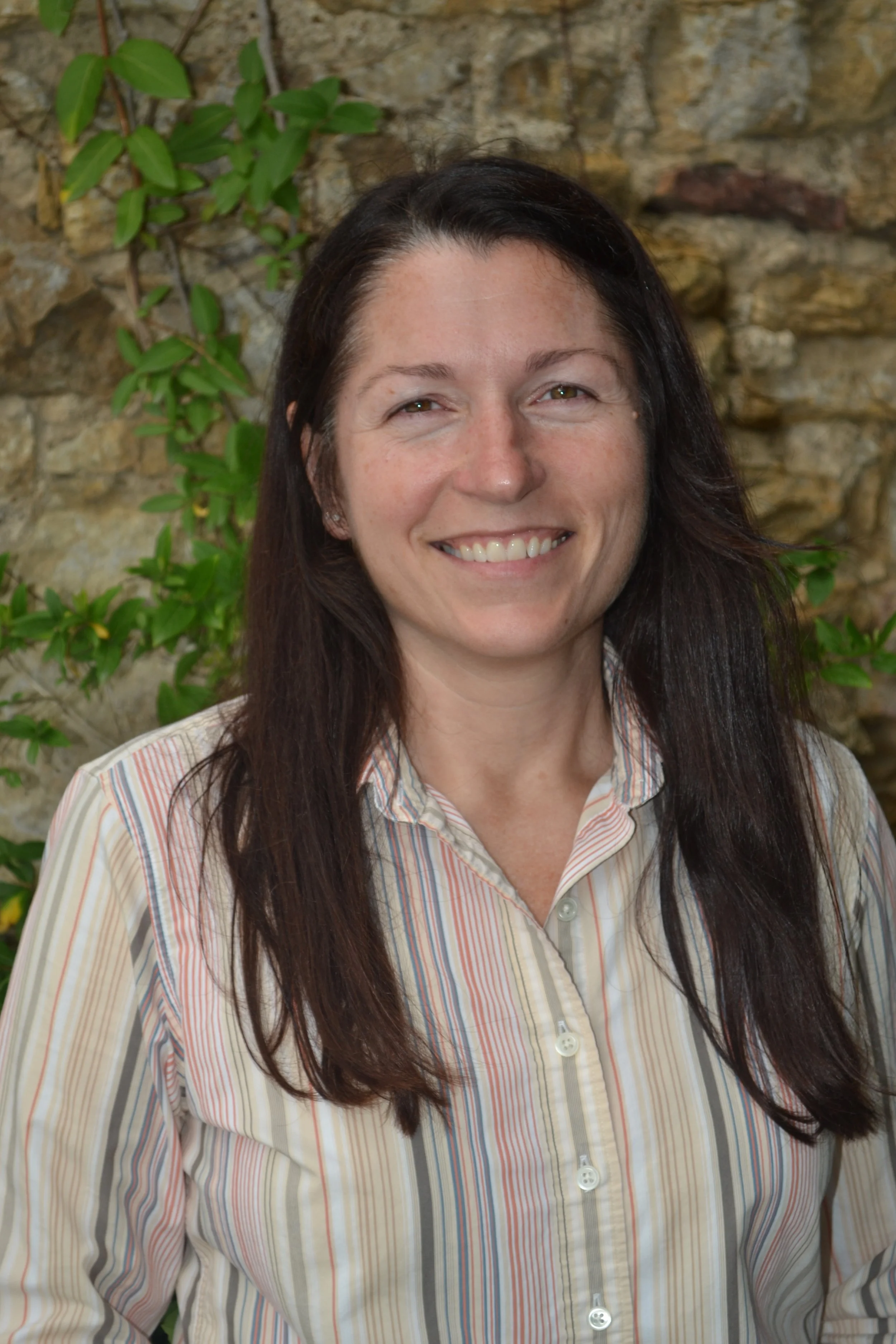 A woman with long dark hair, smiling, wearing a striped shirt, standing in front of a stone wall with green plants.