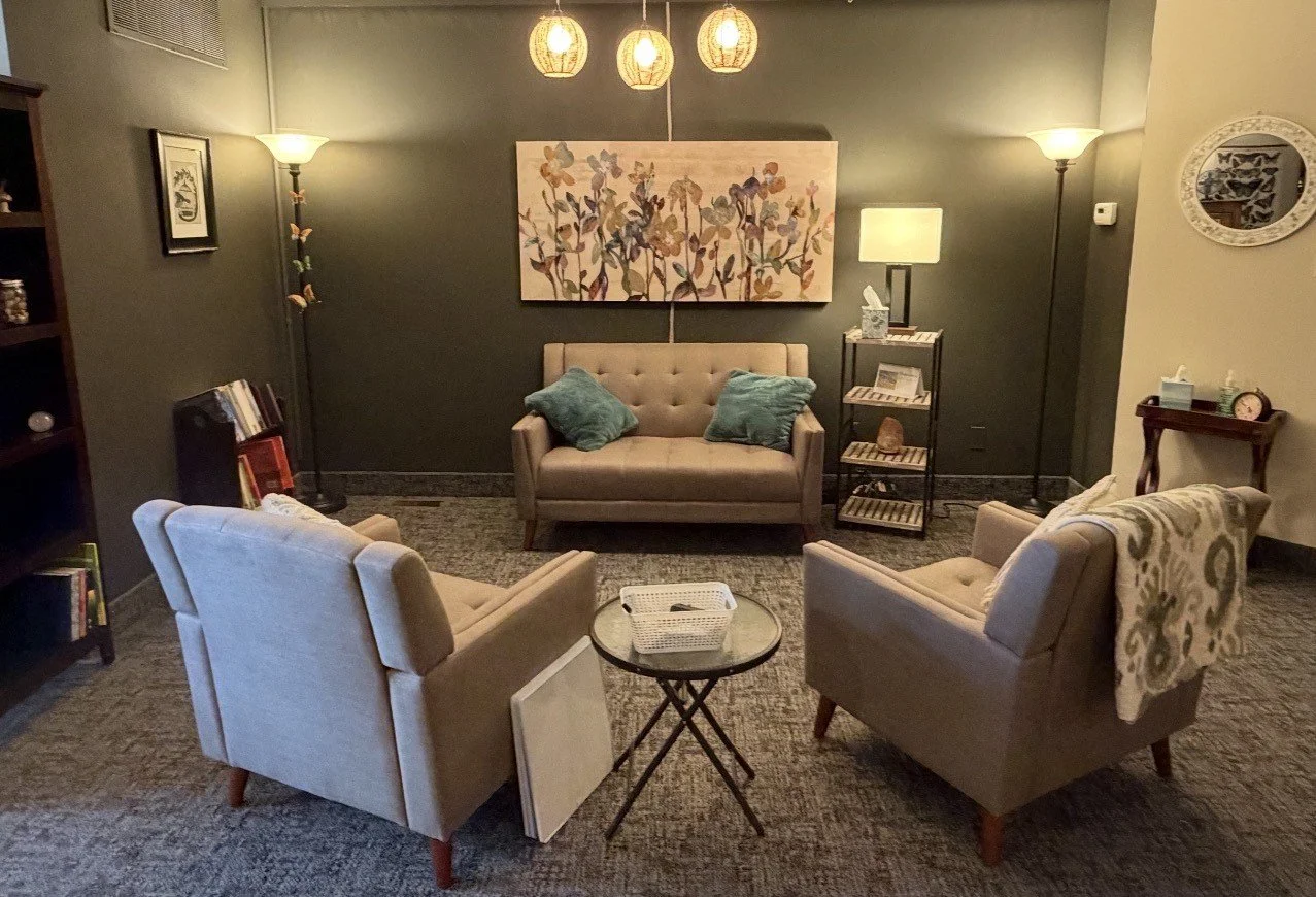 A cozy living room with three beige armchairs arranged around a small round glass table. A beige loveseat with two teal pillows is centered against a dark green wall, which is decorated with a floral painting. There are two tall floor lamps and a bookshelf on the left side, with a small table and clock on the right. The room has warm lighting from three ceiling pendant lights.