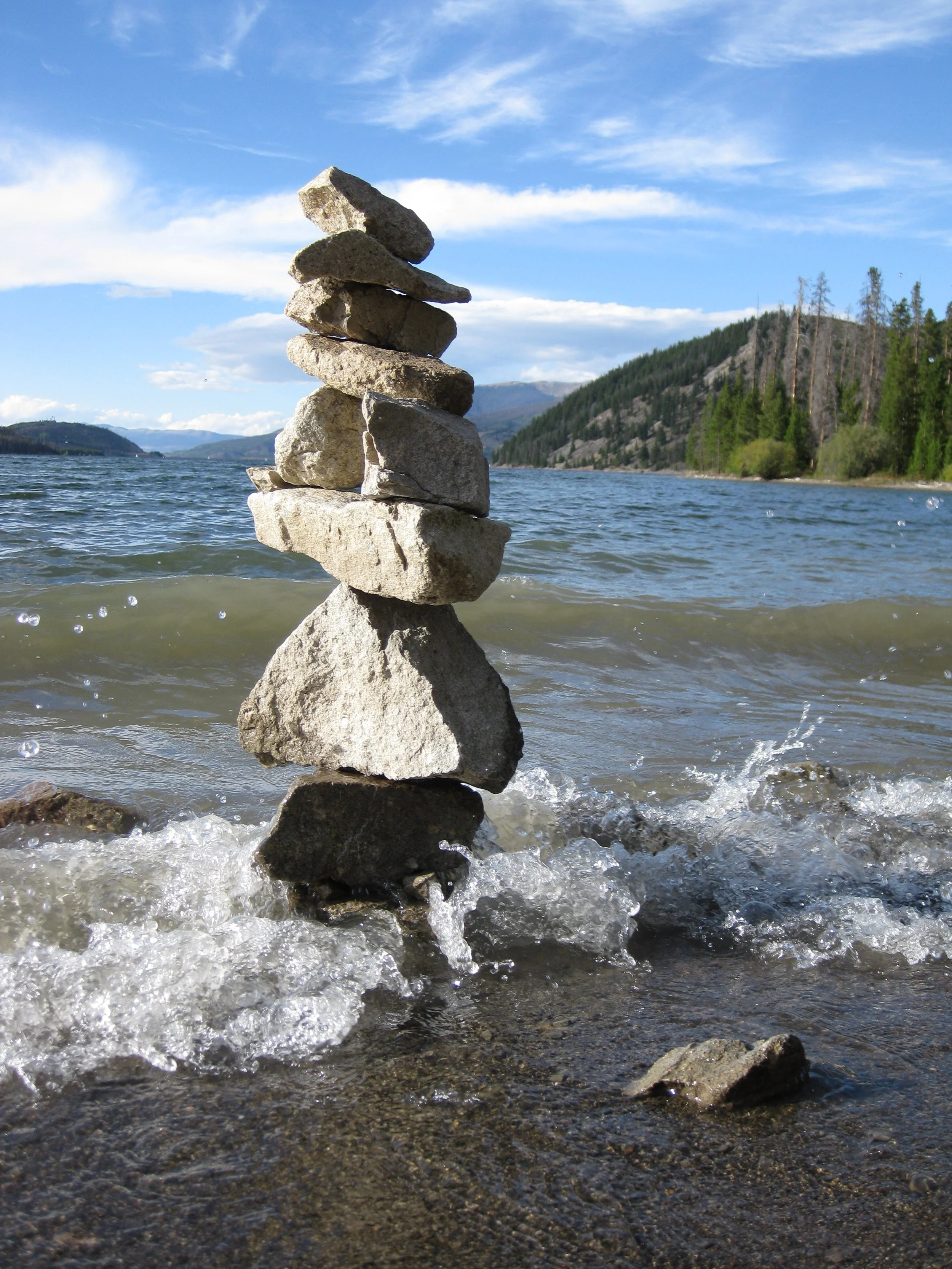 A stack of rocks balanced in the water near a lakeshore, with mountains and a forested hillside in the background under a partly cloudy sky.