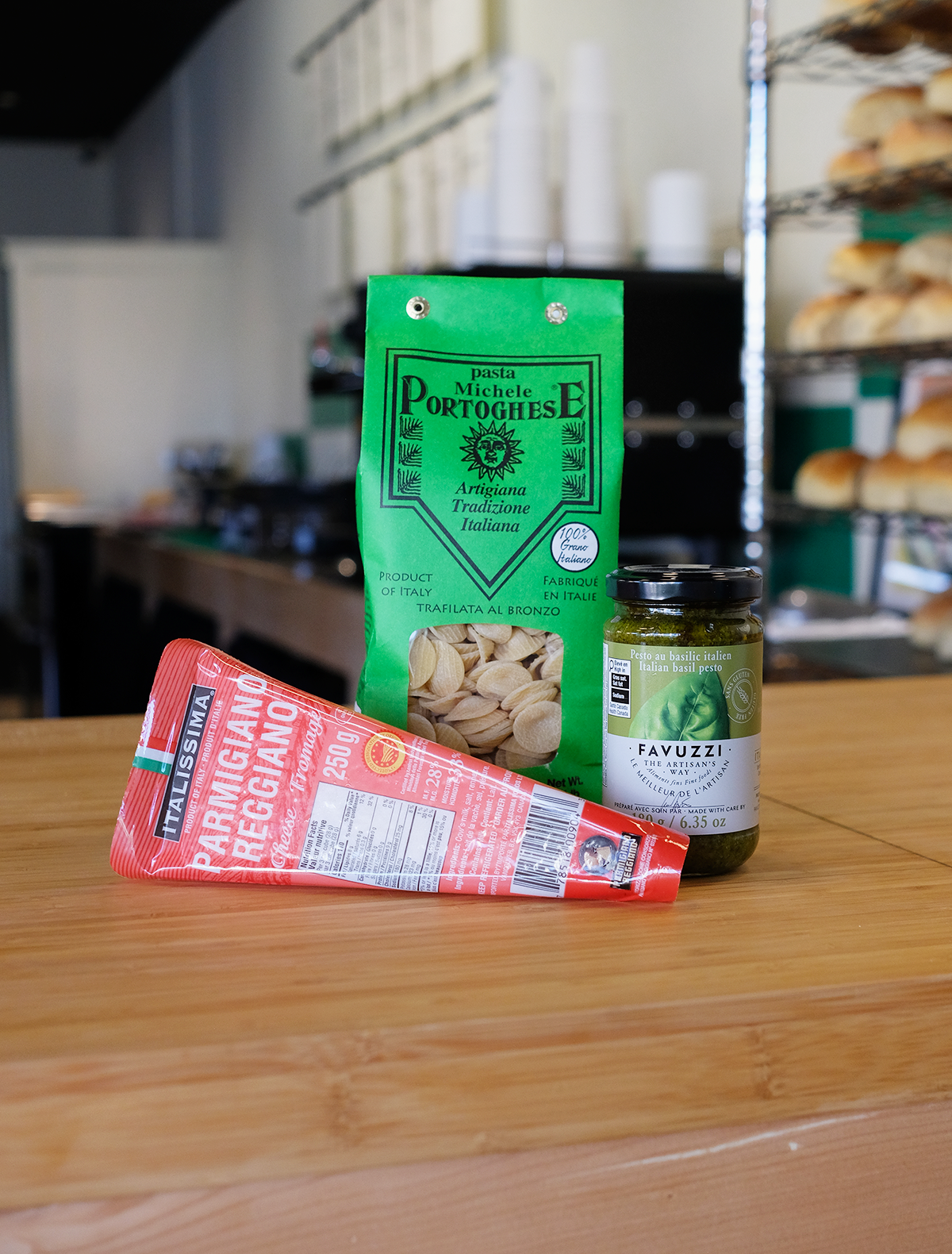A green bag of Michele Portoghese pasta, a jar of Italian basil pesto, and a package of Parmigiano Reggiano cheese on a wooden surface in a kitchen setting.