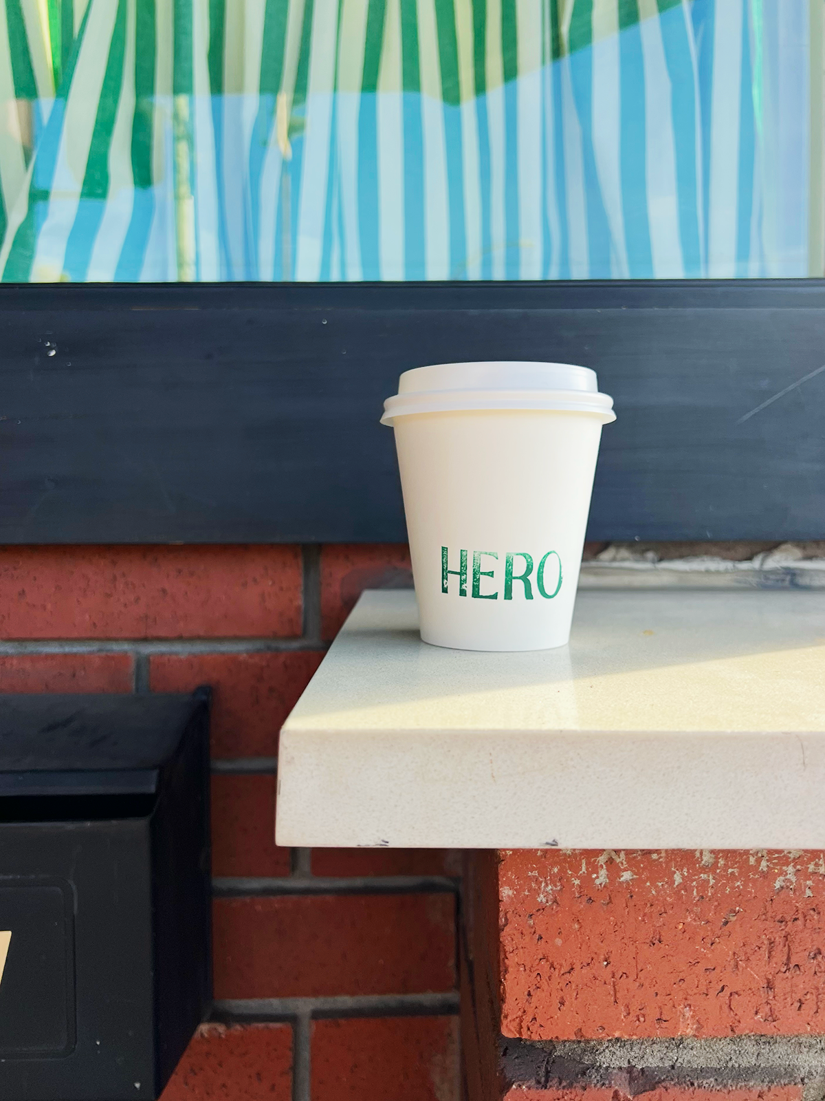 A white paper coffee cup with a plastic lid, sitting on a white table outside. The cup has the word 'HERO' printed on it in green letters. In the background, there is a brick wall and a black object at the corner.