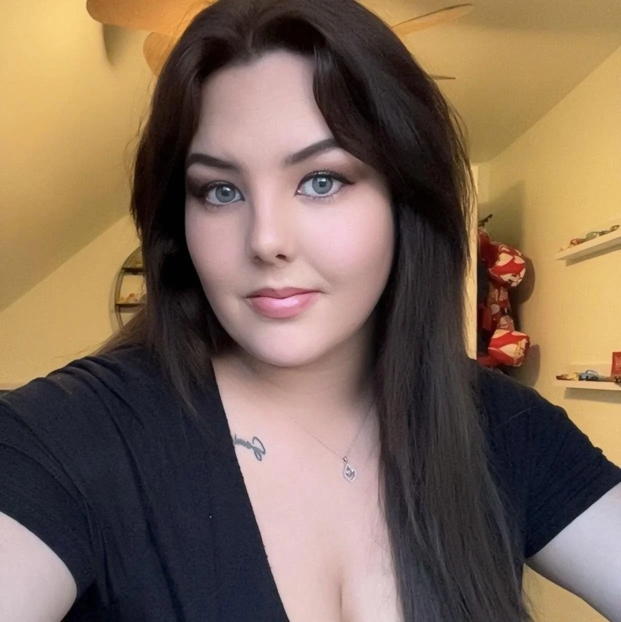 A woman with long dark hair and blue eyes taking a selfie indoors with shelves and red and white objects in the background.