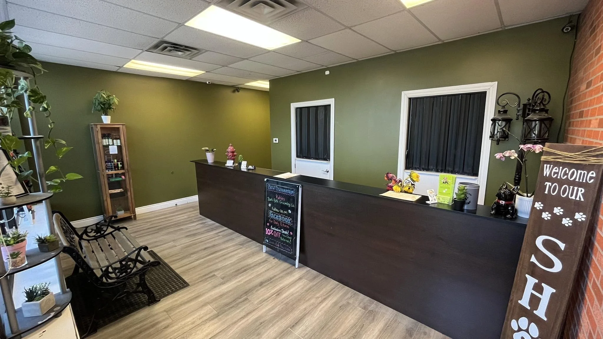 Interior of a pet grooming salon with a dark reception desk, a chalkboard sign, and a waiting area with benches, decorated with plants, lanterns, and a sign that says 'Welcome to our' with the rest cut off.